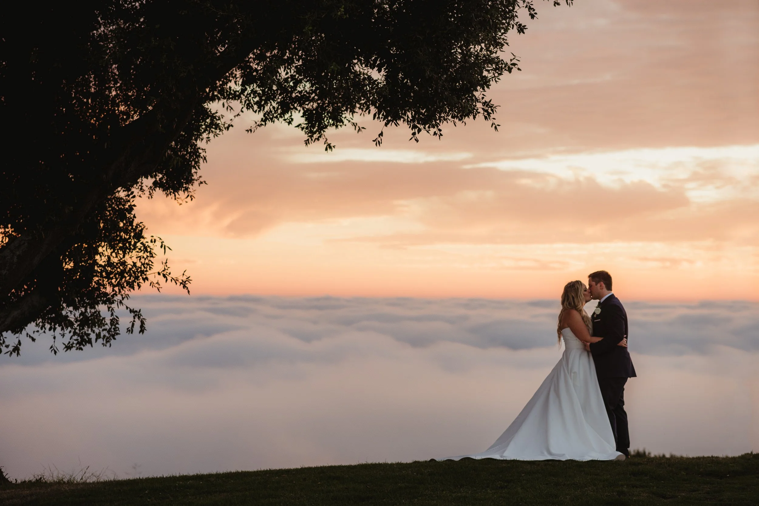 A bride and groom kiss in front of a sunset overlooking a bed of clouds
