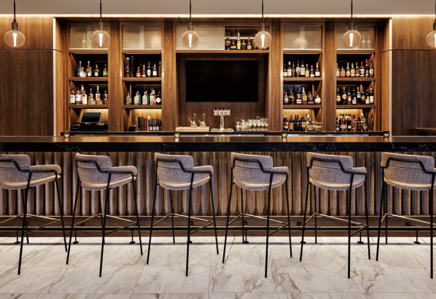 Modern bar with a wooden back wall displaying an array of liquor bottles, a black marble countertop, and six upholstered bar stools with metal legs lined up in front.