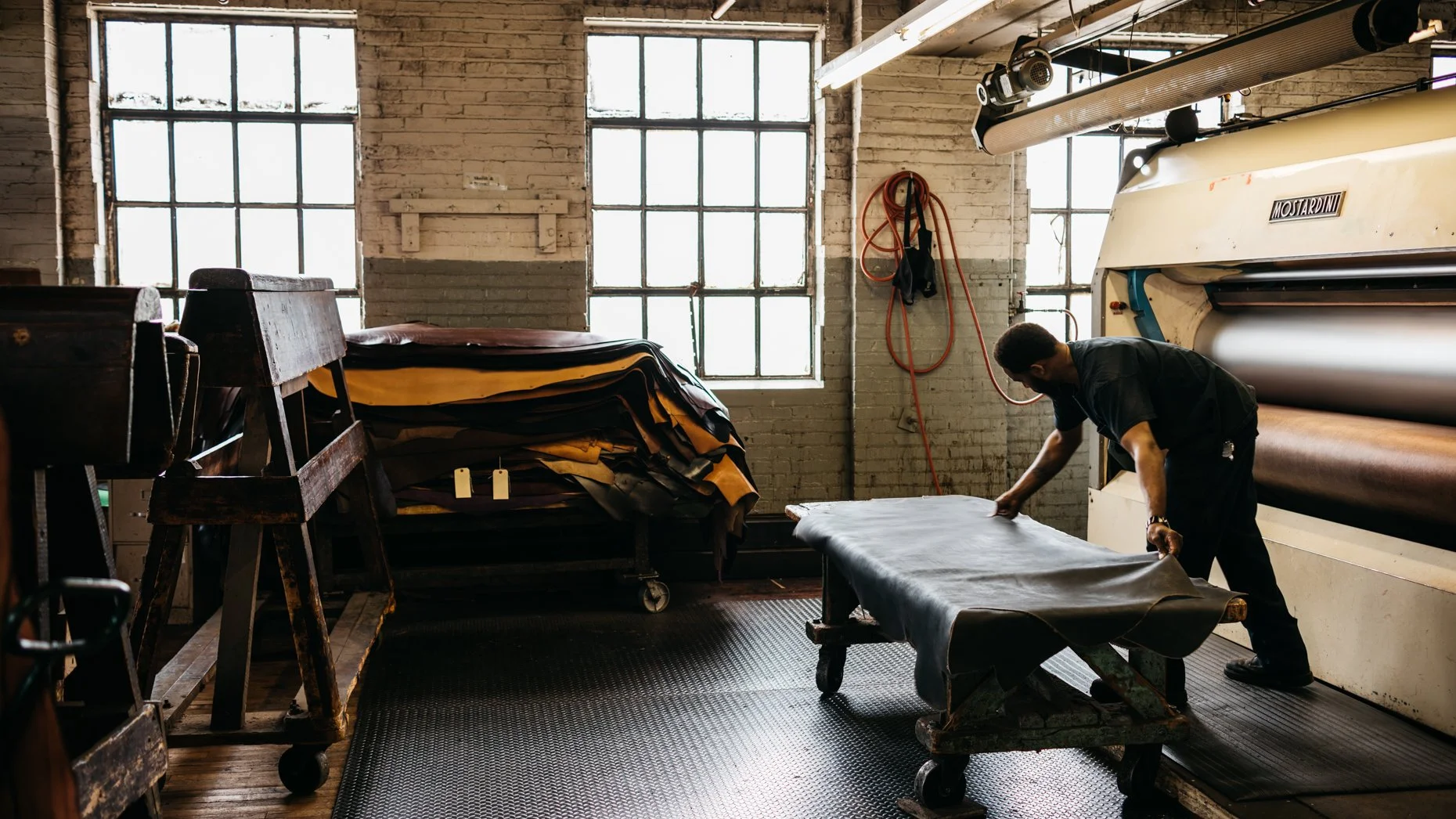 A worker handling leather in a workshop with large windows, leather rolls, and industrial equipment.