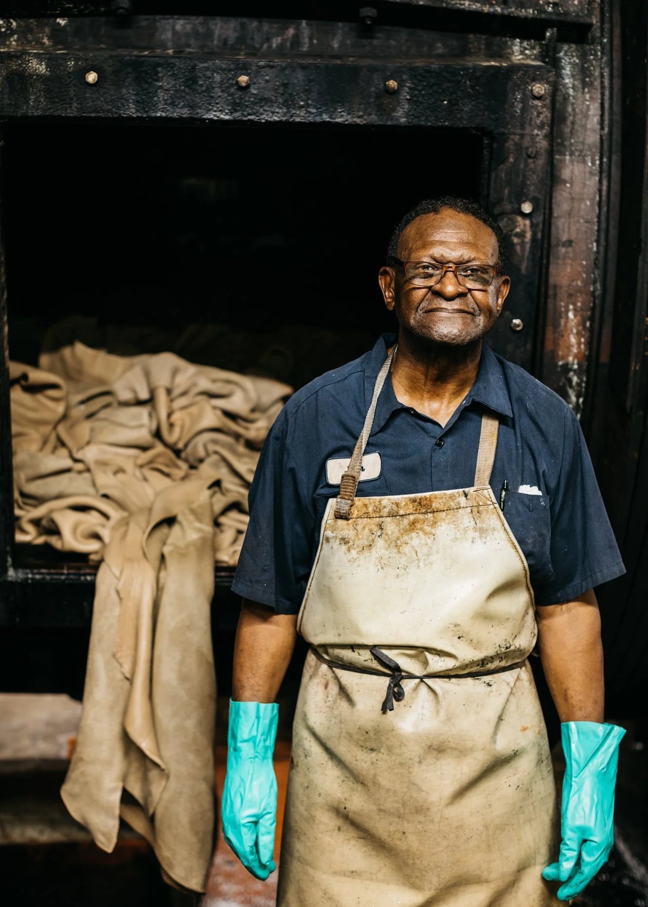 A middle-aged man wearing glasses and a navy blue shirt stands in front of a large tannery tumbler with leather hides inside. He is wearing a stained apron and blue rubber gloves, and has a serious expression.