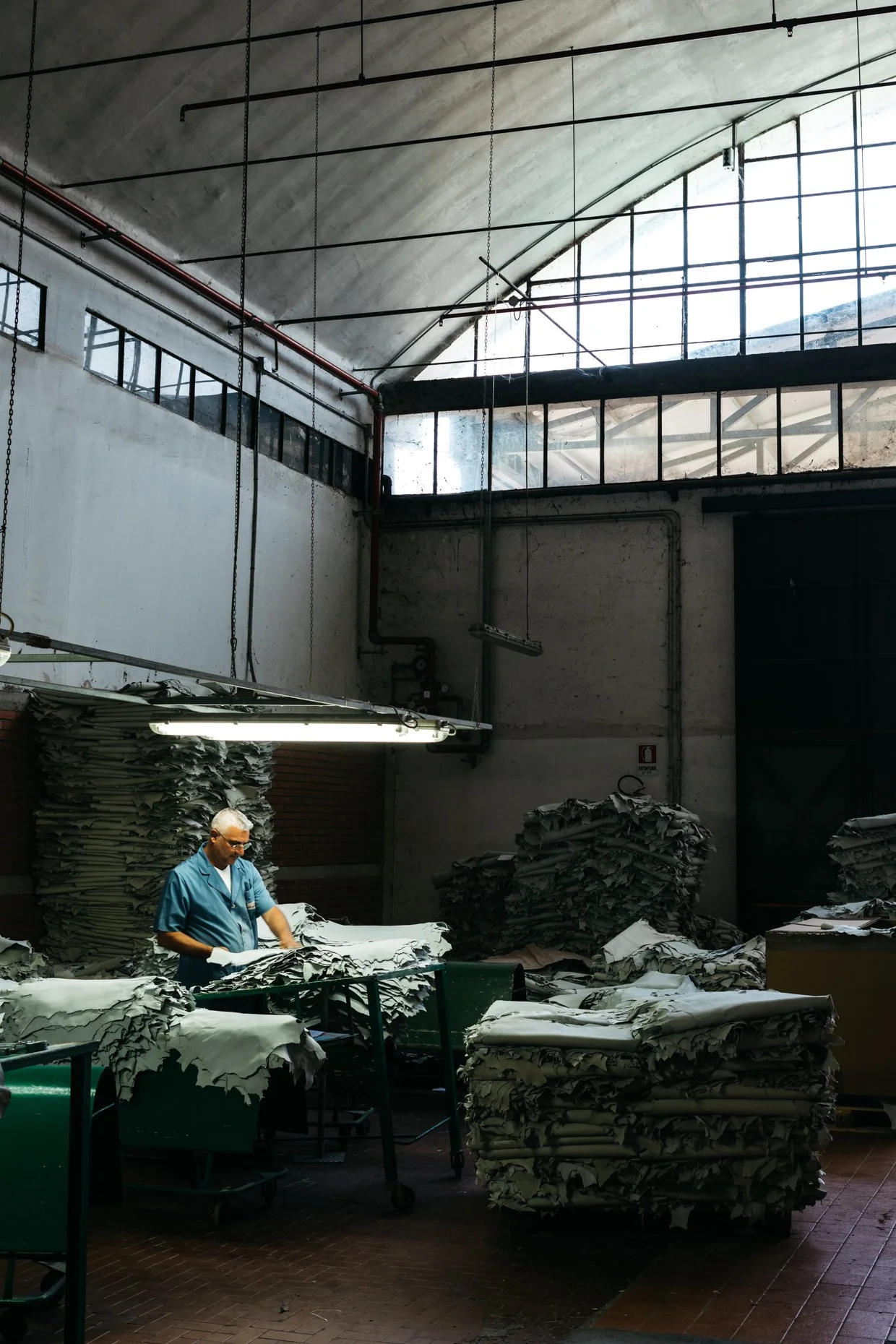 Worker examining stacks of leather in an tannery with high ceilings and large windows.