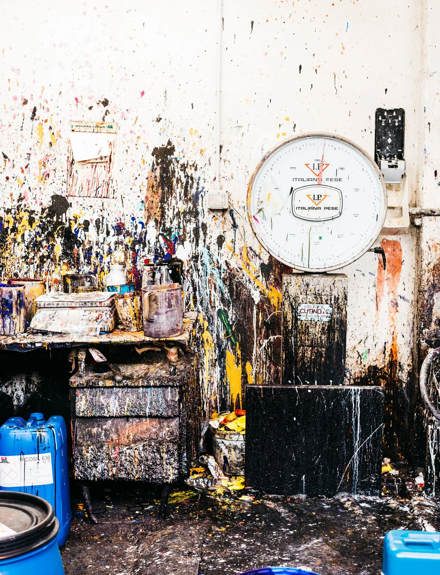 Dye-splattered tannery room with wall clock, dye cans, brushes, and a cluttered work surface.