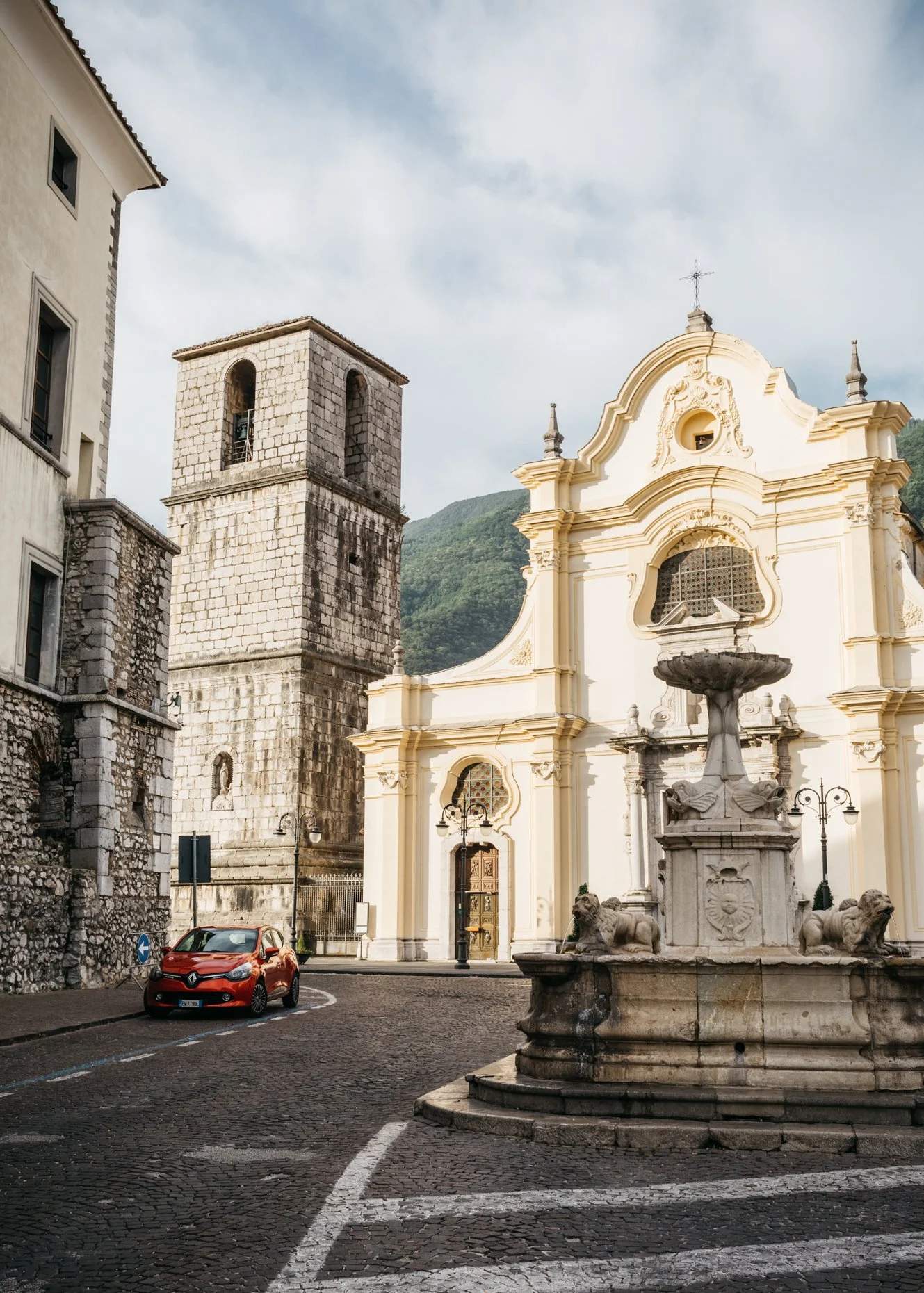 A narrow cobblestone street with an orange car parked near a historic white church, featuring a fountain with lion sculptures in the foreground and a stone tower on the left, with mountains in the background under a partly cloudy sky.