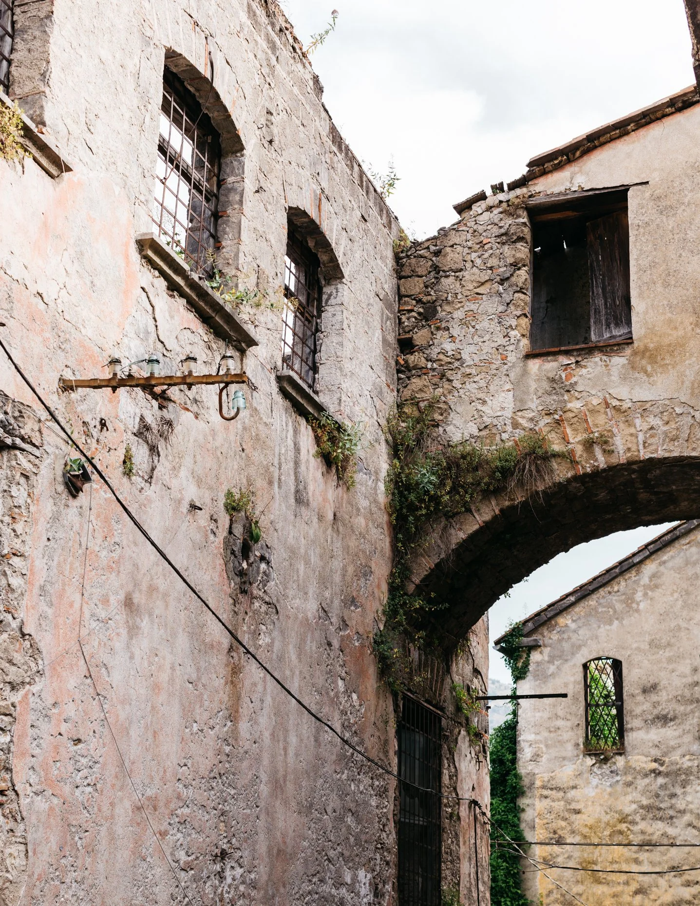 A narrow alley between old stone buildings, with weathered walls, small barred windows, and an arched stone bridge connecting the structures. Faint greenery grows on the walls.