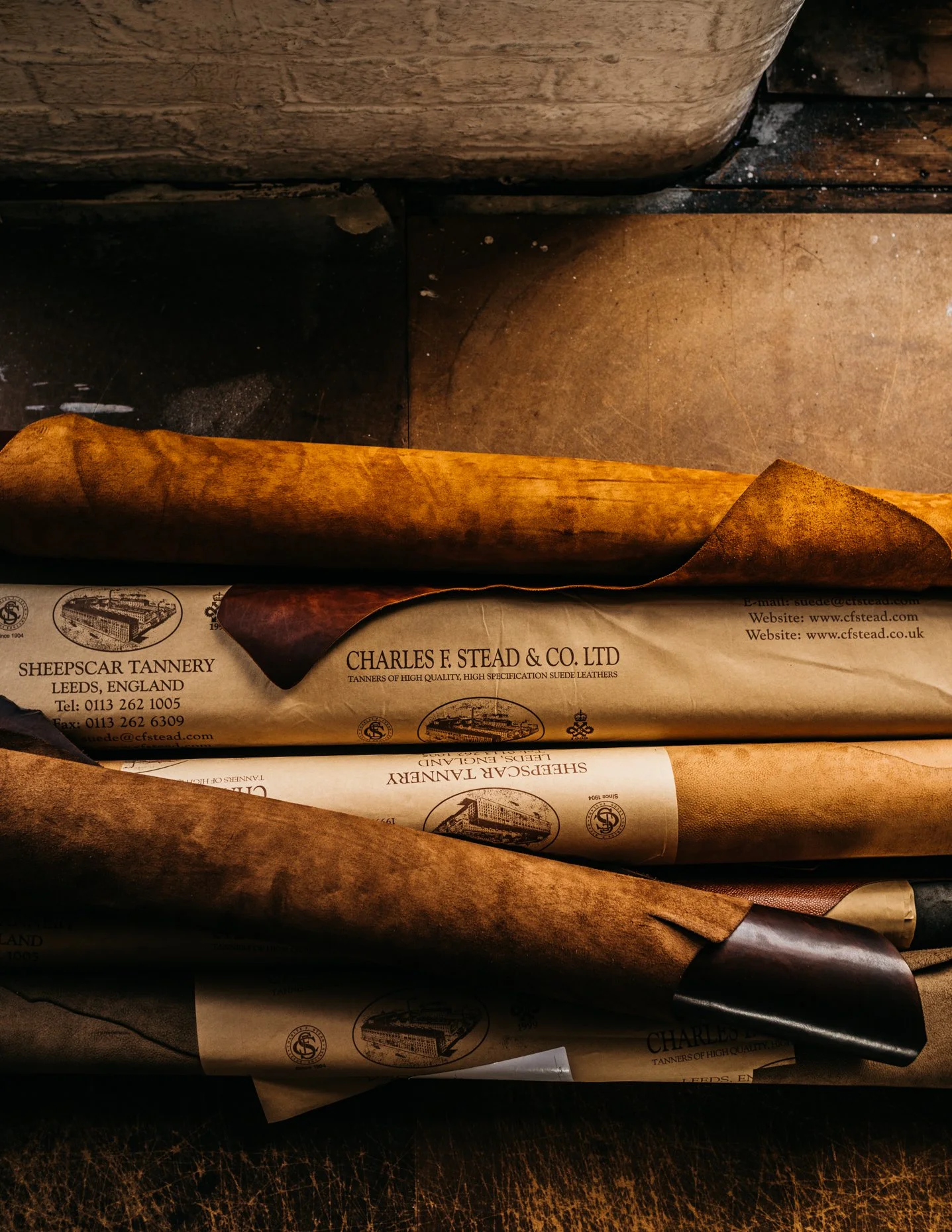 Rolled leather hides wrapped in paper with branding from Charles F. Stead & Co Ltd, a tannery in Leeds, England, on a wooden surface.