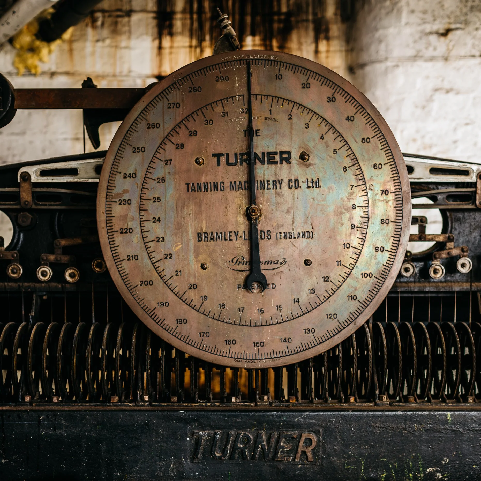 A vintage weighing scale with a circular dial, displaying the brand name 'Turner', used by Tanning Machinery Co. Ltd. and Bramley-Lords, England, placed on an old machine.