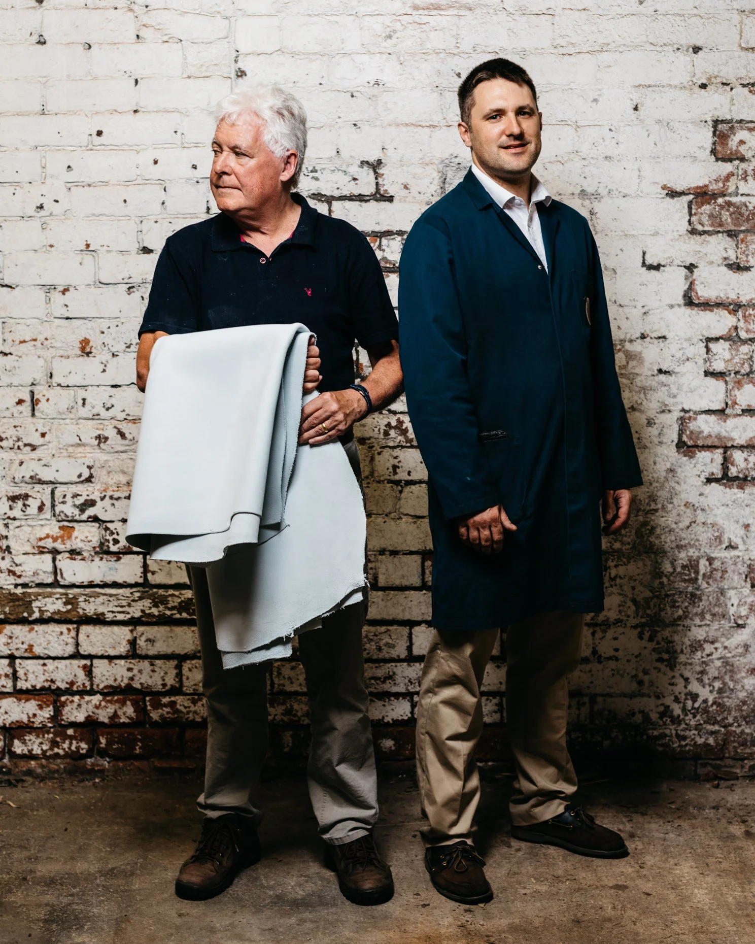 Two men standing in a tannery against a white brick wall, one holding leather hides and the other wearing a workcoat.