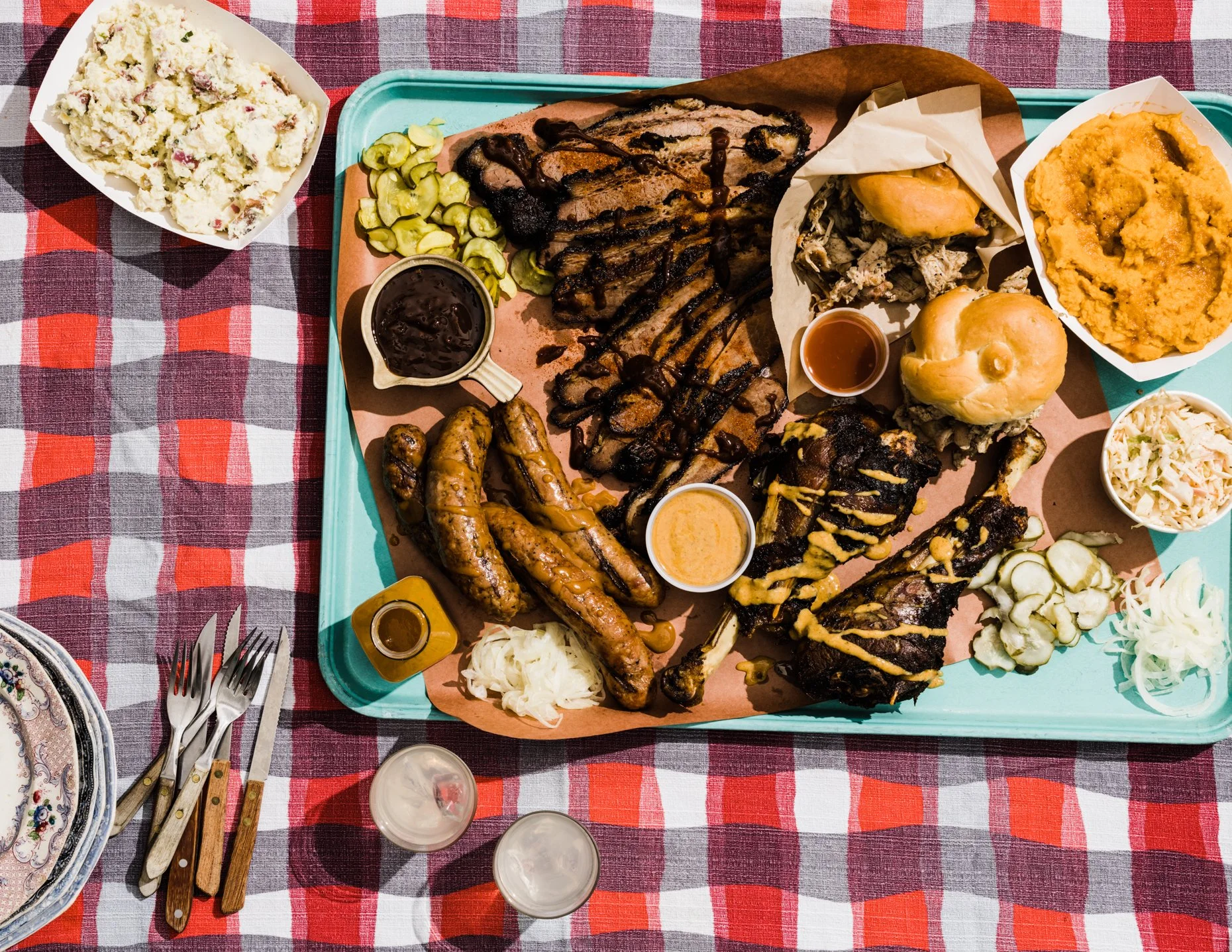 A barbecue platter with ribs, grilled chicken, sausage links, baked beans, potato salad, coleslaw, pickles, onions, and buns on a blue tray. There is potato salad in a white bowl, and a small container of barbecue sauce on a red and black checkered t
