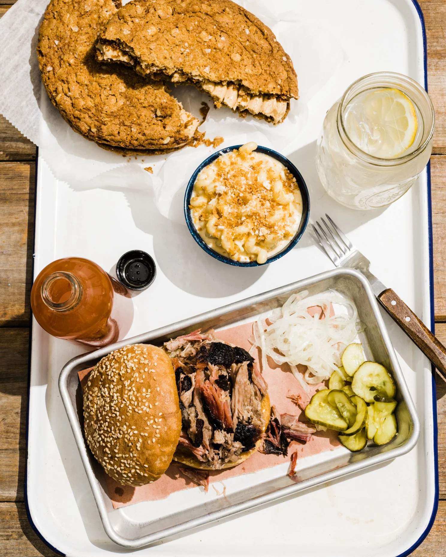 Tray with a sesame seed bun hamburger, sliced pickles and onions, shredded meat, a bowl of macaroni and cheese, a cookie with white chocolate chips, a mason jar of lemonade with lemon slices, and a bottle of hot sauce.