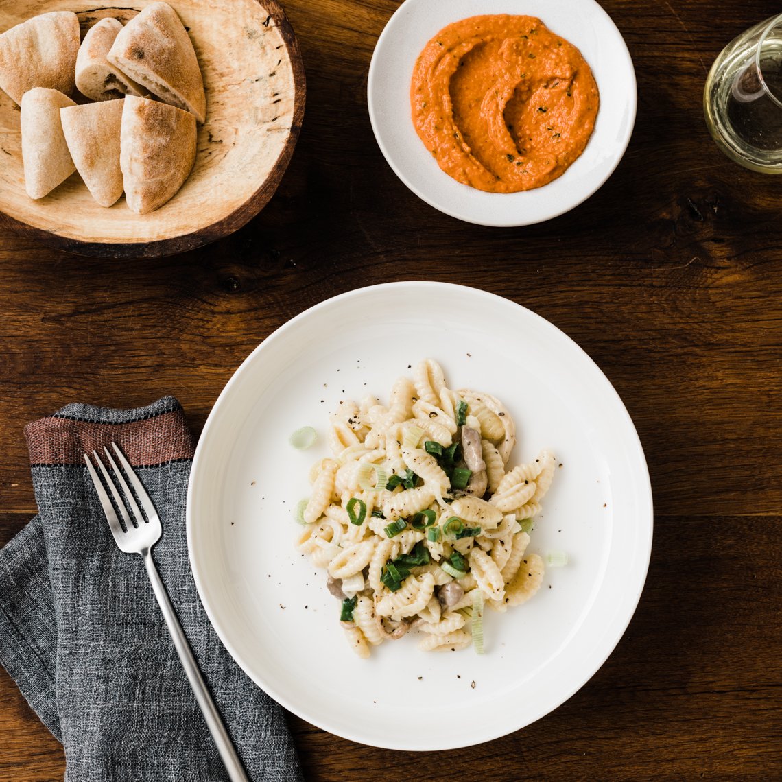 A plate of cooked pasta garnished with green onions, a bowl of red sauce, a wooden plate of pita bread, and a glass of white wine on a dark wooden table.