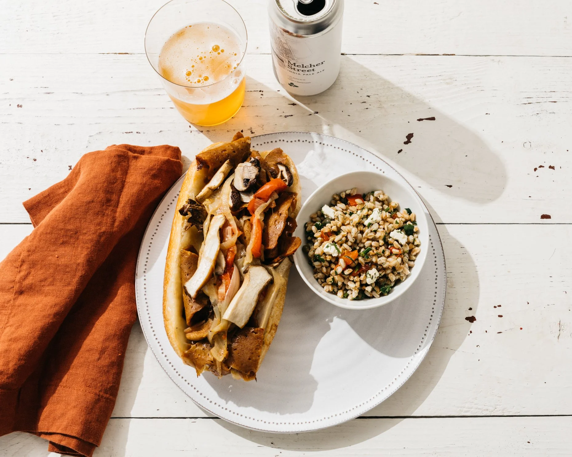 A plate with a hot dog topped with grilled mushrooms, onions, and peppers, a side bowl of chopped grain salad with vegetables and cheese, a can of beer, and a glass of beer on a white wooden table with an orange cloth napkin.