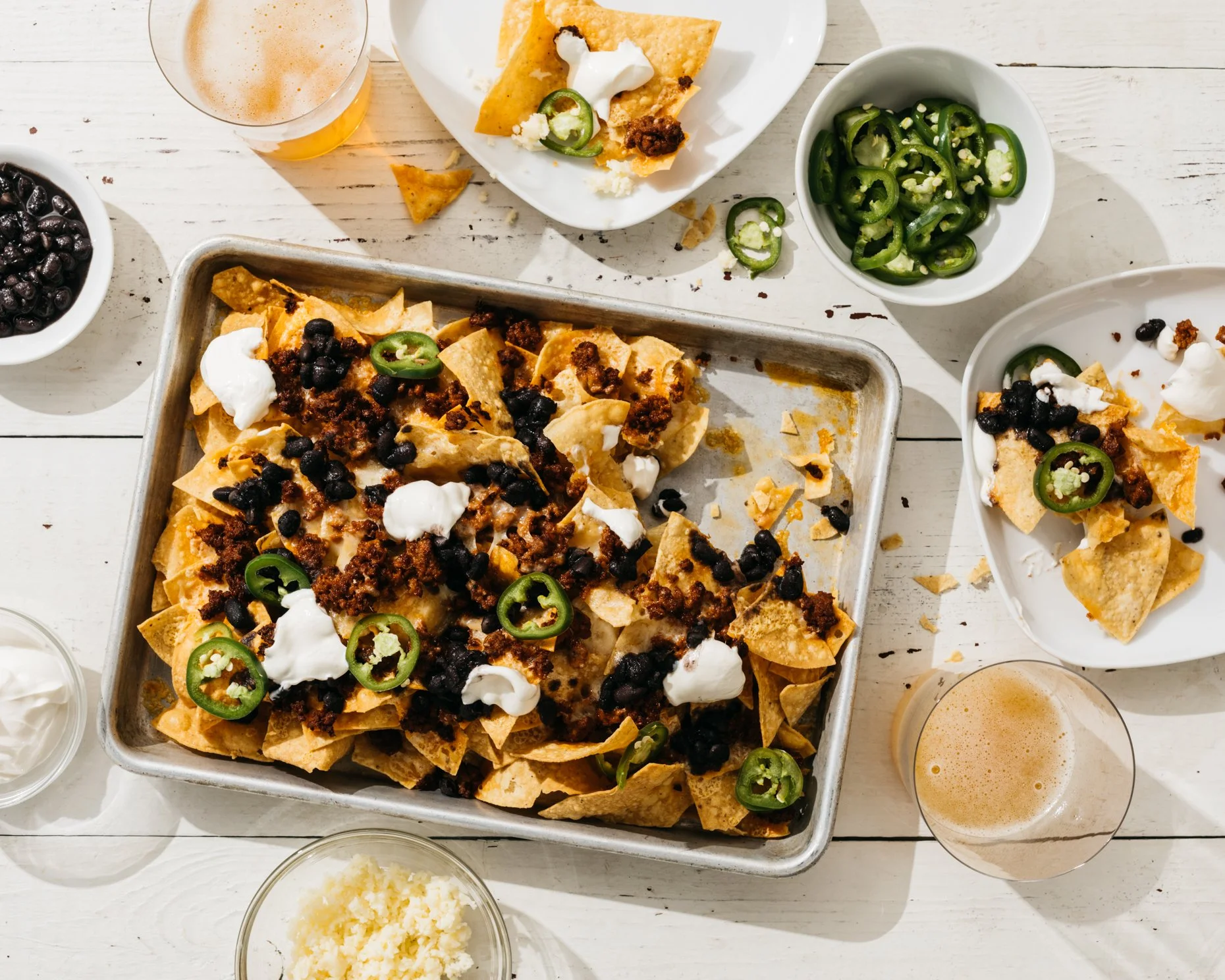 Nachos topped with black beans, sour cream, jalapeños, and ground beef, with bowls of jalapeños, black beans, cheese, and beer around it.