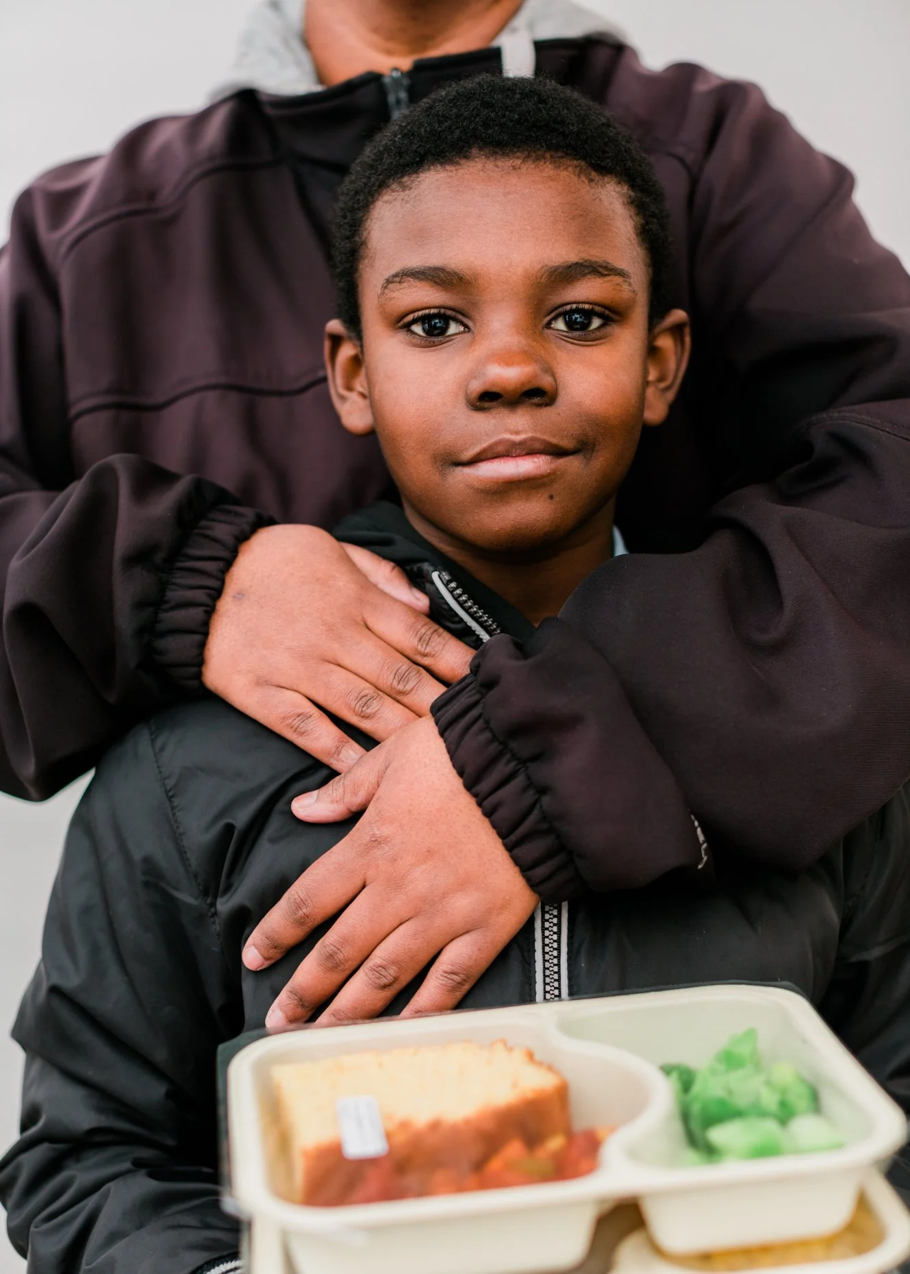 A young boy with short black hair and dark skin sits with an adult standing behind him, who has their arms around the boy. The boy is wearing a black jacket, and there is a beige lunch tray in front of him containing a piece of cake and some green ve