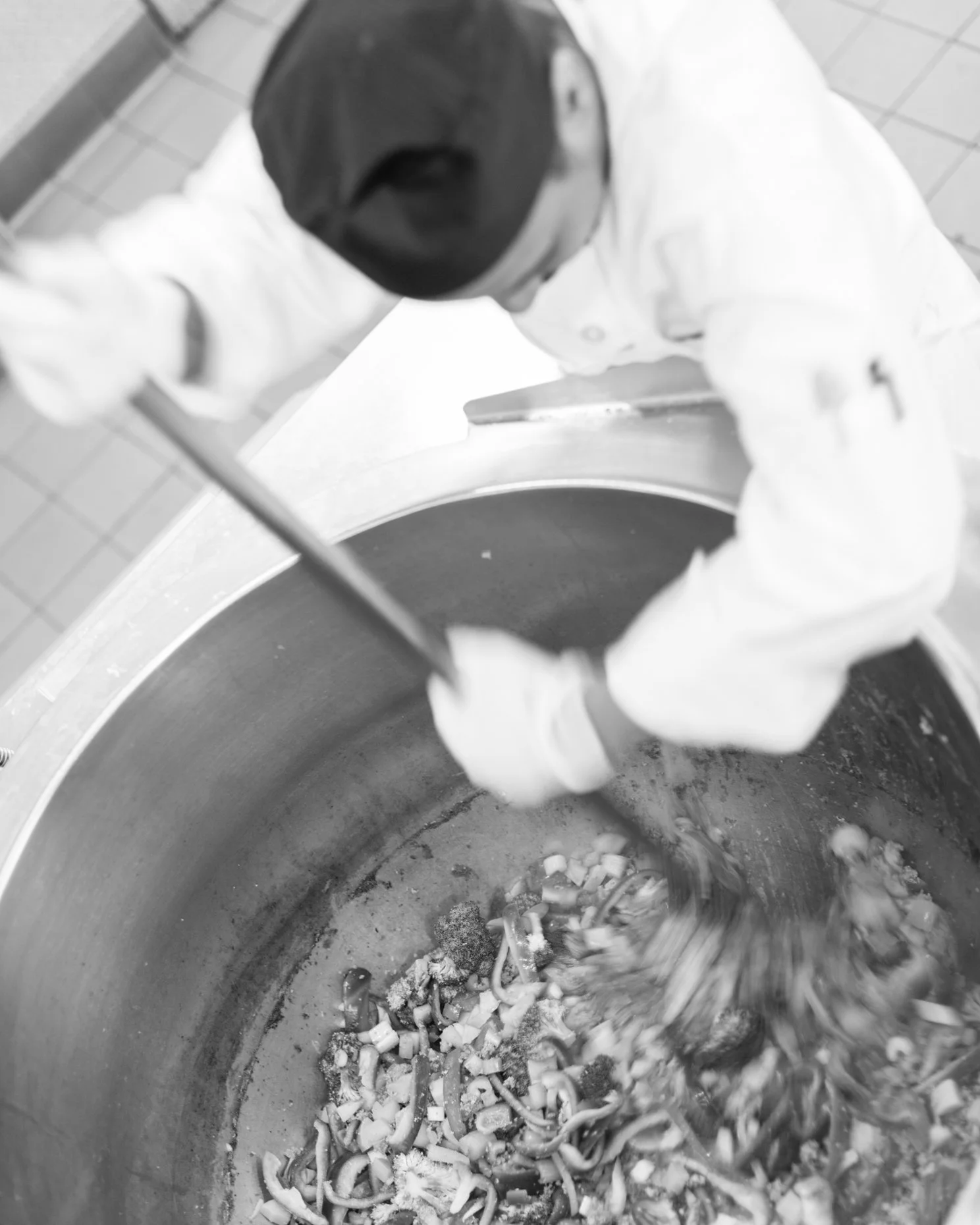 A chef wearing a black cap, white coat, and gloves stirring a large pot of cooked vegetables in a commercial kitchen.