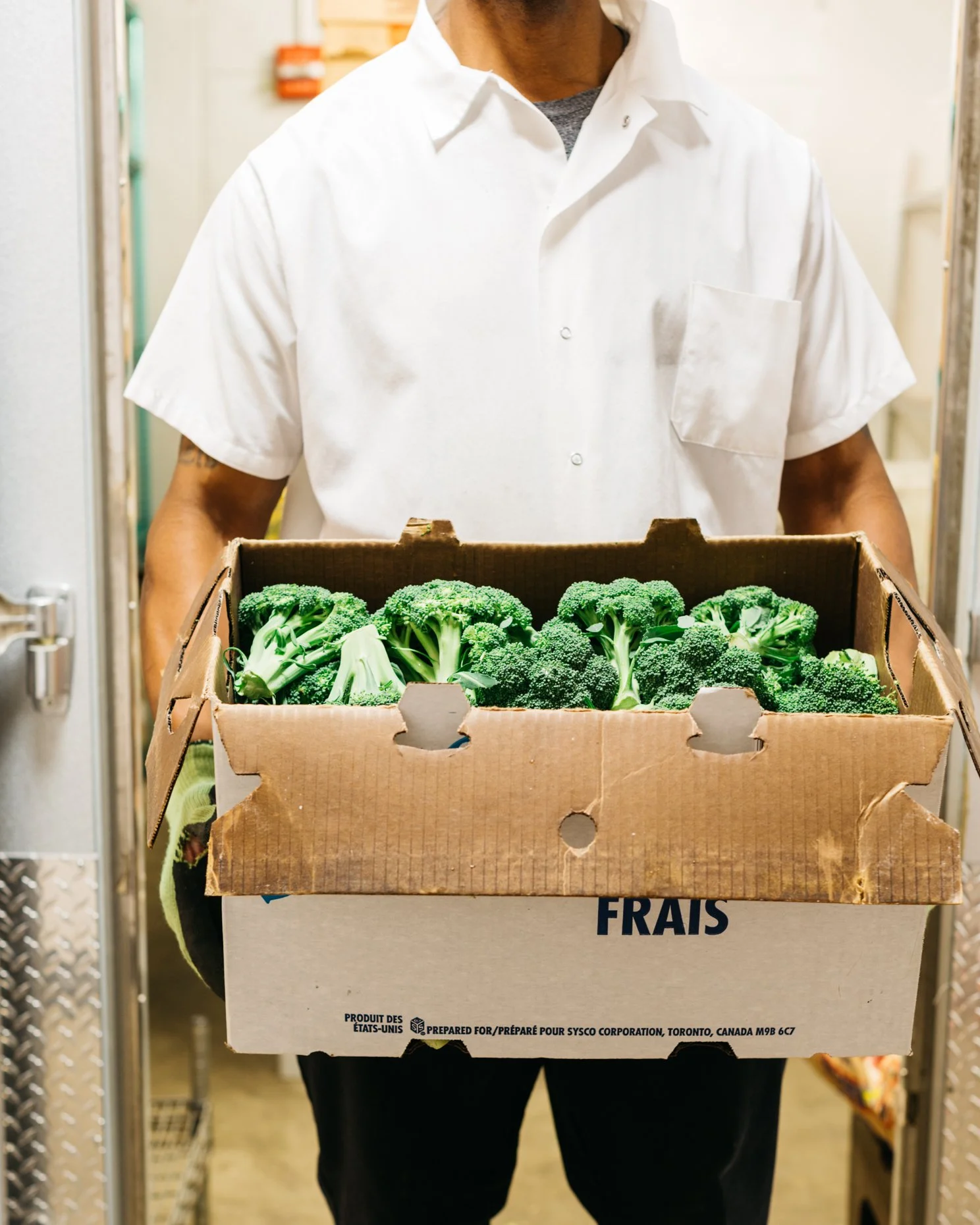 A person wearing a white shirt holding a box of fresh broccoli in a storage or warehouse setting.