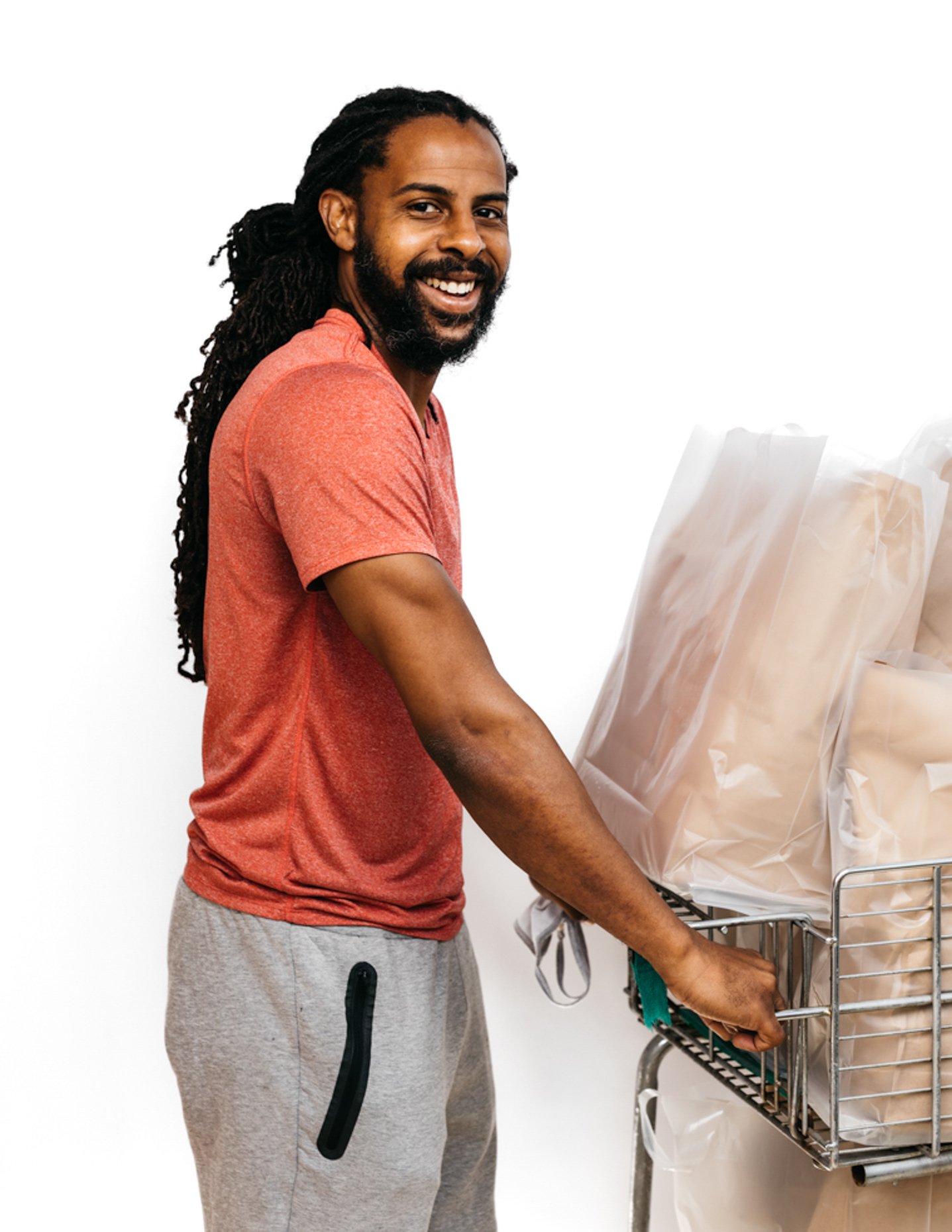 A smiling man with dreadlocks and a beard standing next to a shopping cart filled with paper bags.