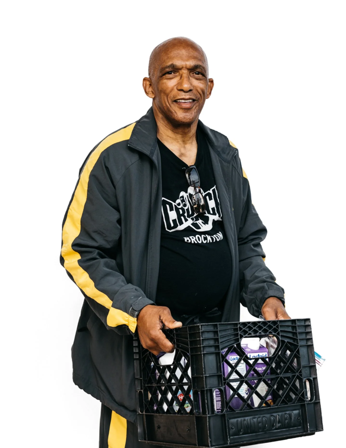 Older man with a graze smile holding a black crate filled with groceries, wearing a black jacket with yellow accents and sunglasses hanging from his shirt.