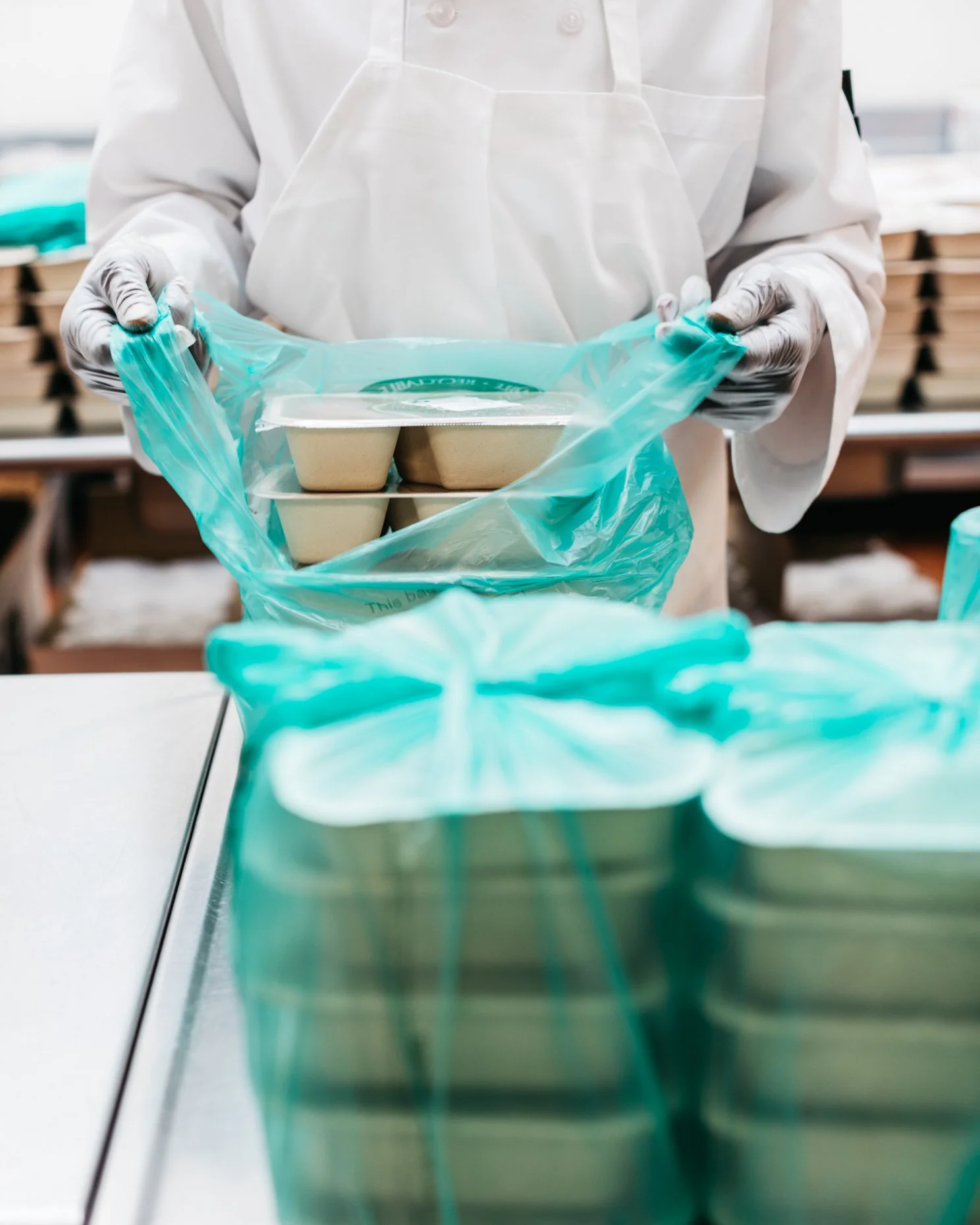 A person in white protective clothing and gloves is placing foam food containers into a teal plastic bag in a commercial kitchen or food processing facility.