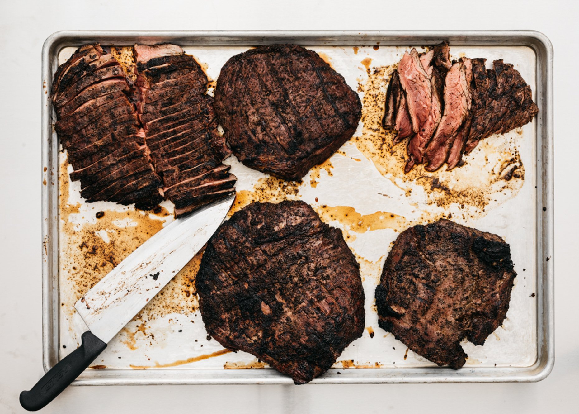 Steaks on a baking sheet,  and sliced steak, with a carving knife.