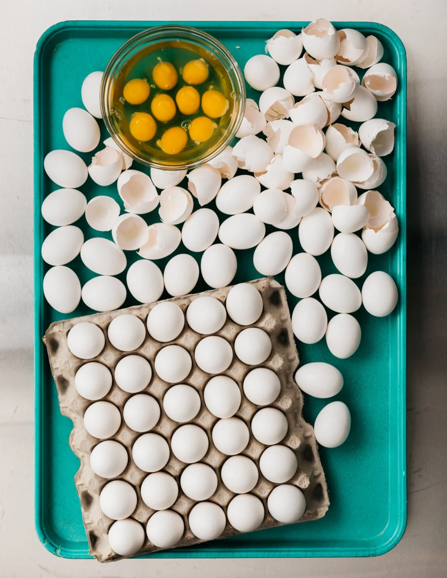 Carton of eggs, cracked eggs in a bowl, and broken eggshells on a teal tray.