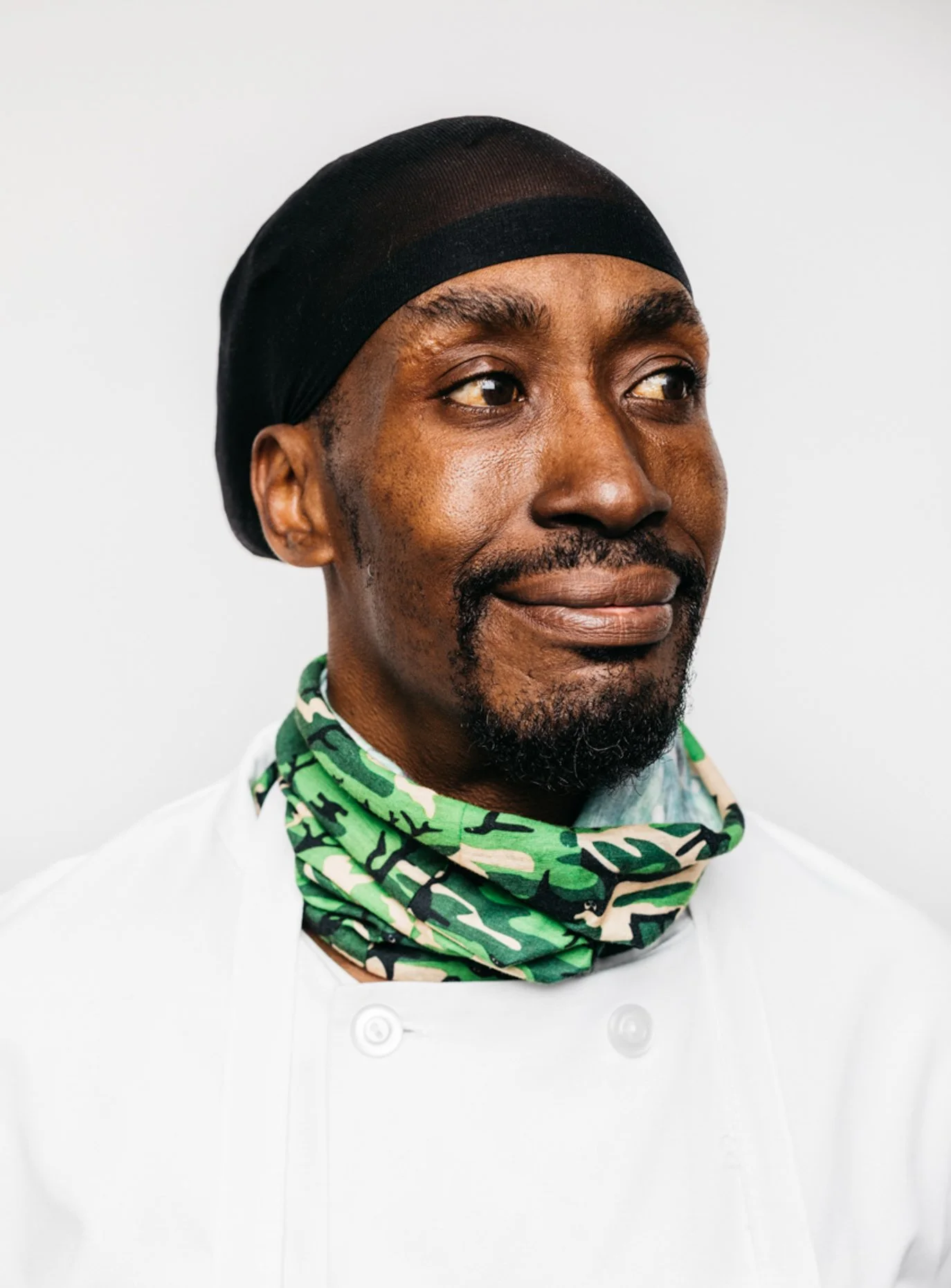 A chef wearing a black hairnet, white chef coat, and a green camouflage neck gaiter, standing against a plain white background.