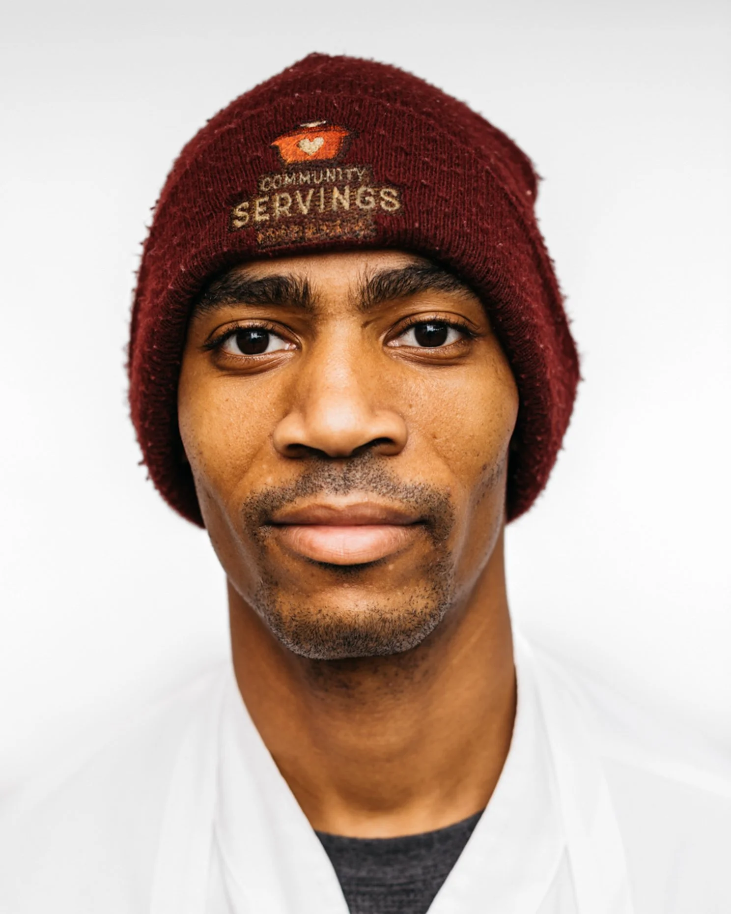 Close-up of a man wearing a maroon beanie with the words 'Community Servings' embroidered on it, against a plain white background.