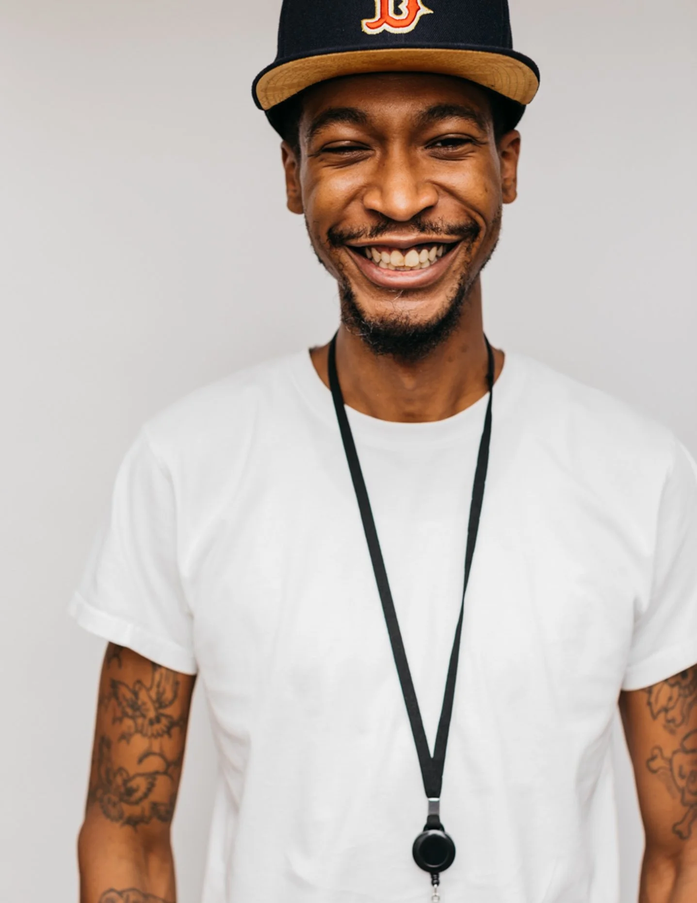 Smiling man wearing a black and yellow baseball cap, white T-shirt, and a lanyard with a black whistle, standing against a plain background.