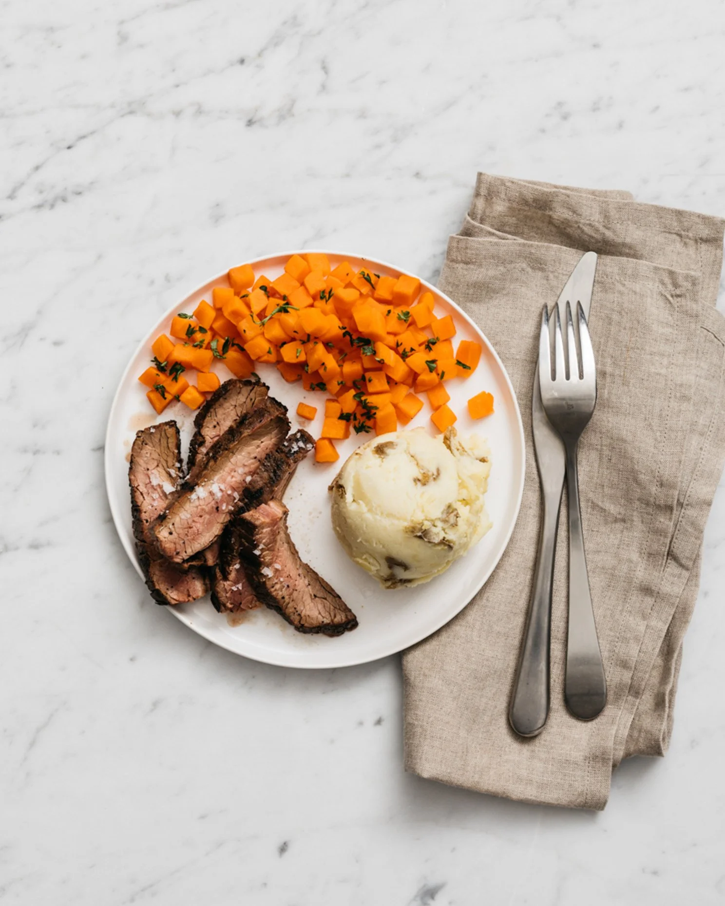 Plate with sliced steak, diced carrots garnished with herbs, and mashed potatoes on a marble surface with fork and knife wrapped in a beige napkin.