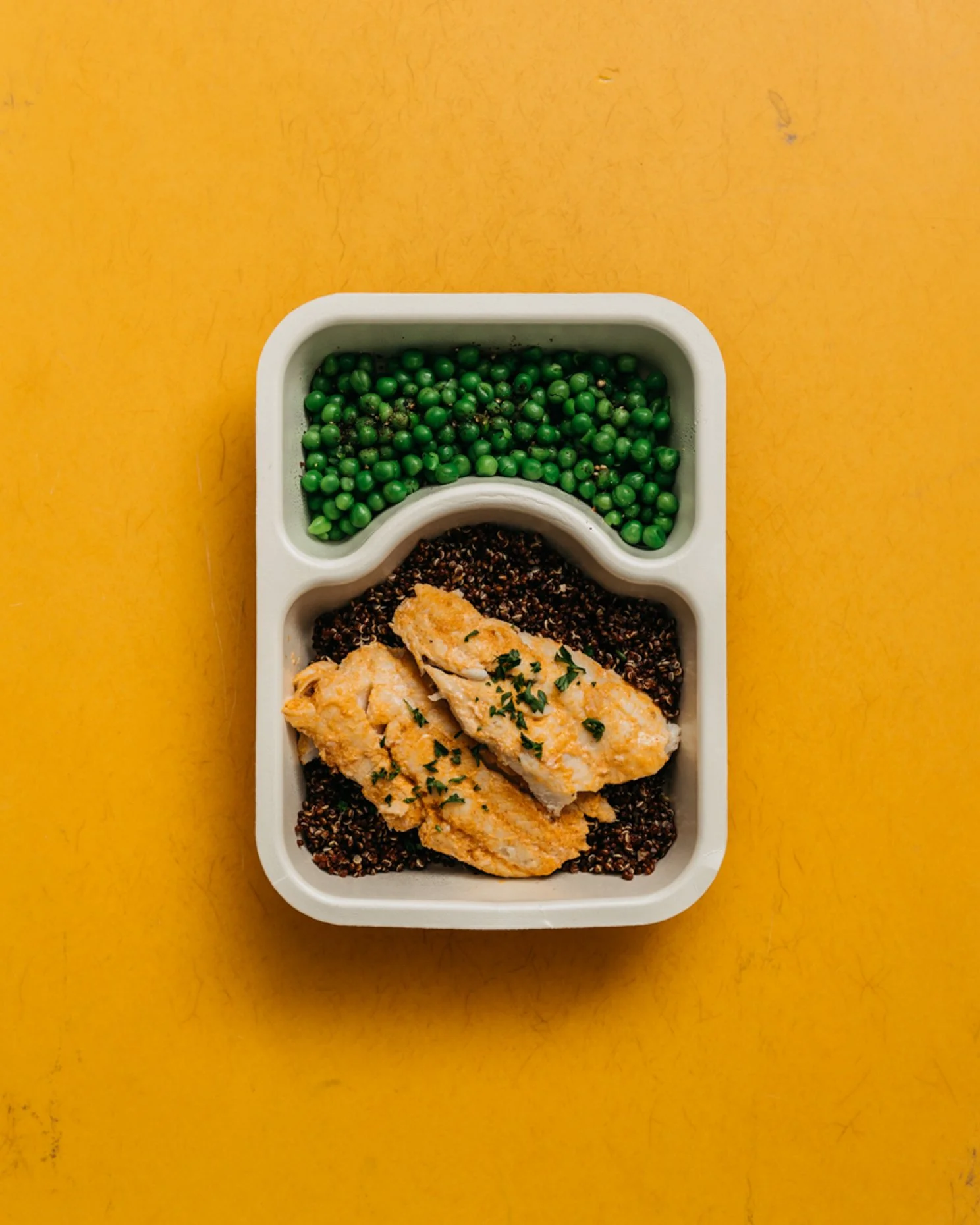A white segmented meal tray with cooked chicken breast garnished with herbs, cooked quinoa, and green peas, on a yellow surface.