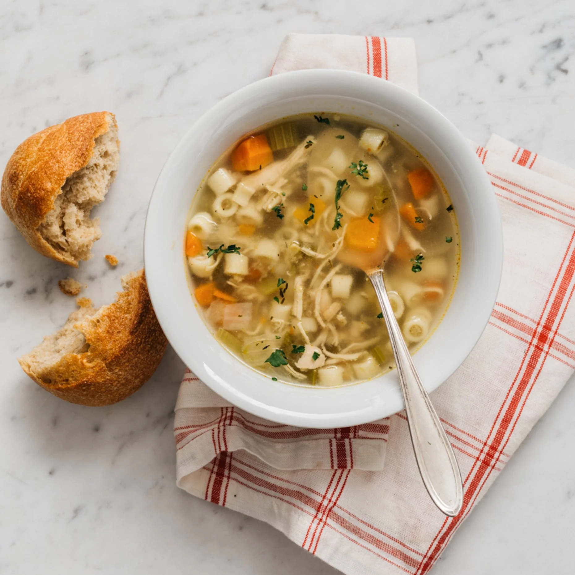 A bowl of chicken noodle soup with diced carrots, celery, and pasta served on a white marble surface, with a piece of bread on the side and a spoon in the soup.
