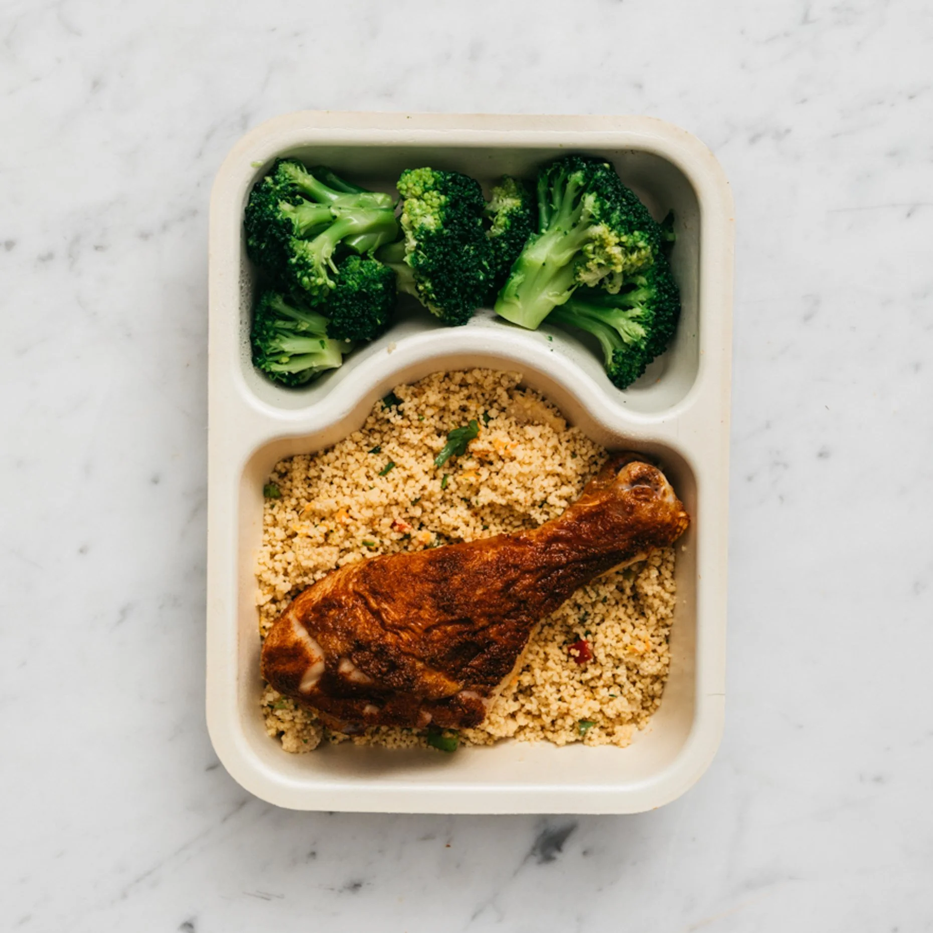 Meal in a compartmentalized container with broccoli, cooked couscous, and a baked chicken leg on a white marble surface.