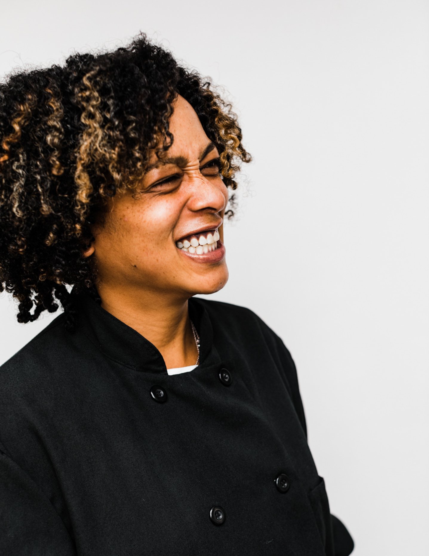 Woman with curly hair smiling and looking to the side, wearing a black chef's coat against a plain white background.