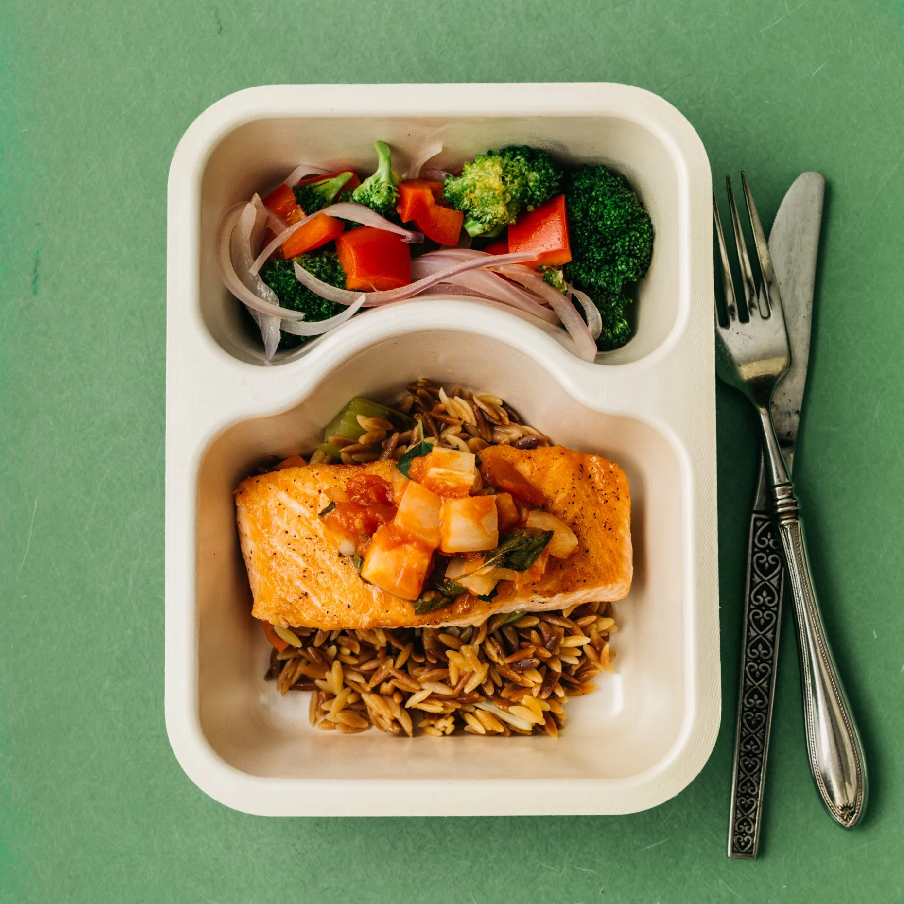 A divided meal tray with cooked broccoli and sliced carrots on the top section, a piece of cooked salmon with vegetables and grains on the bottom section, a fork and knife next to the tray, on a green surface.