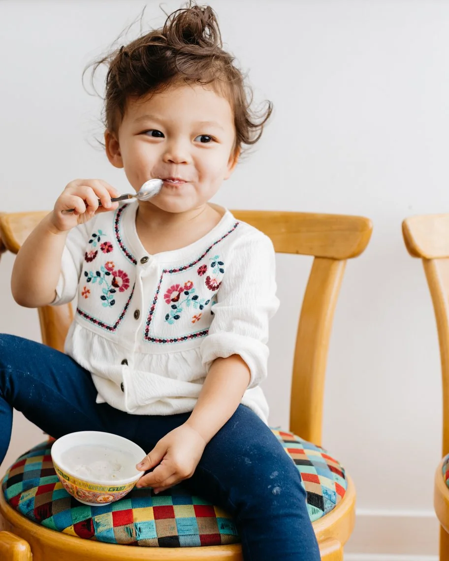 A young child with curly hair is sitting on a wooden chair, eating from a small bowl with a spoon, wearing a white embroidered blouse and dark pants.