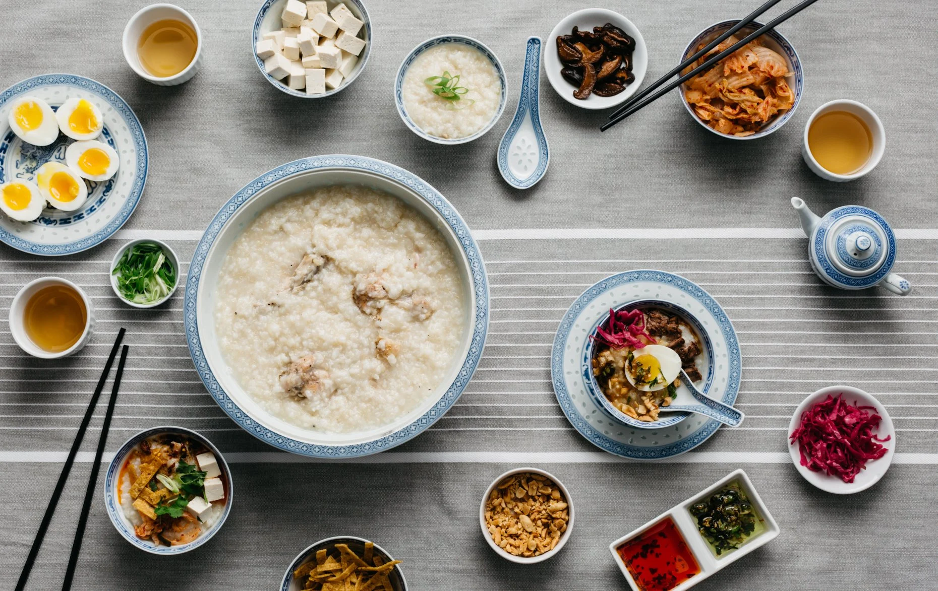 A traditional Asian meal spread on a table, including congee, miso, side dishes, bowls of tea, and garnishes, served on blue and white porcelain dishes and bowls.