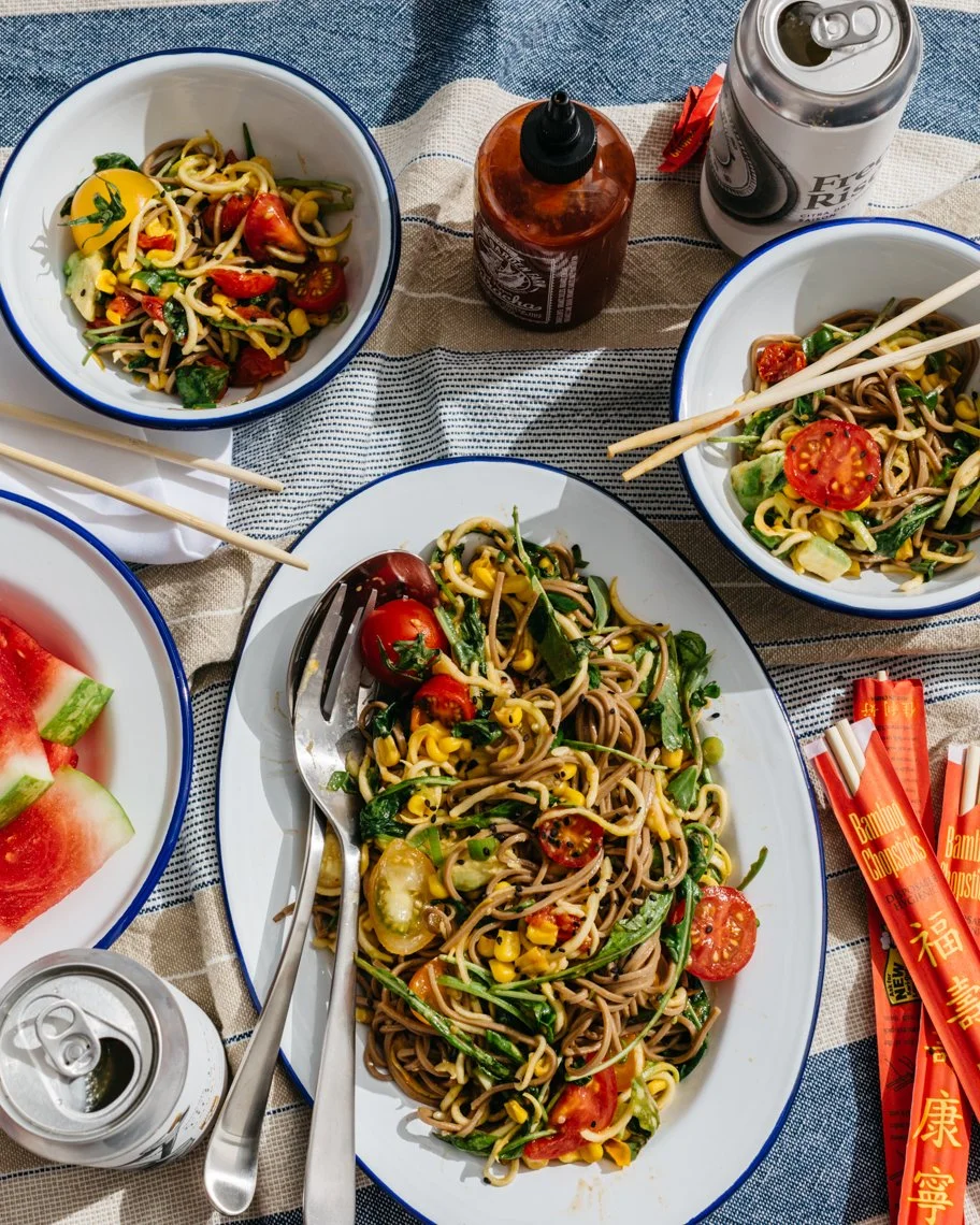 Plated Asian-style noodle salad with cherry tomatoes, chopped vegetables, and chopsticks, on a striped tablecloth with drinks and watermelon slices.