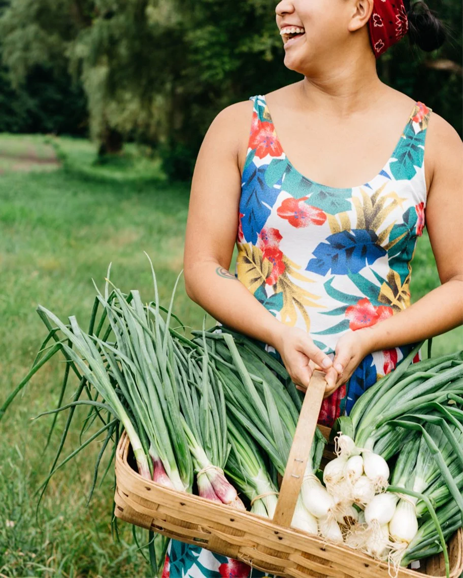 A smiling person with a red bandana holding a basket of fresh green onions and garlic outdoors in a grassy area.