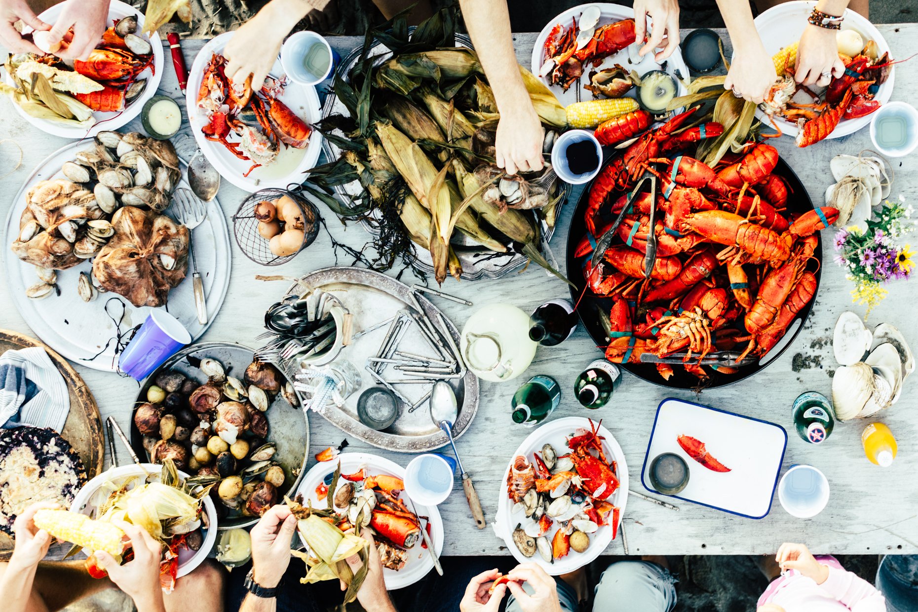 A top-down view of a seafood feast with lobsters, crawfish, shellfish, corn, onions, and vegetables on a table, with people serving themselves.