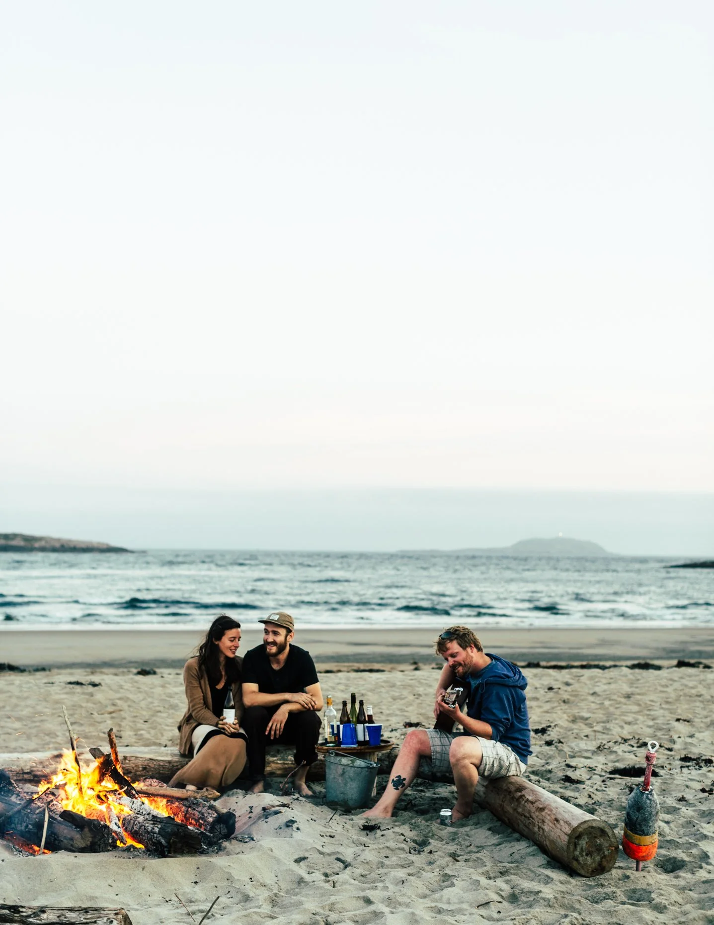 Three people sitting on a beach around a campfire, enjoying drinks and music, with the ocean and sky in the background.
