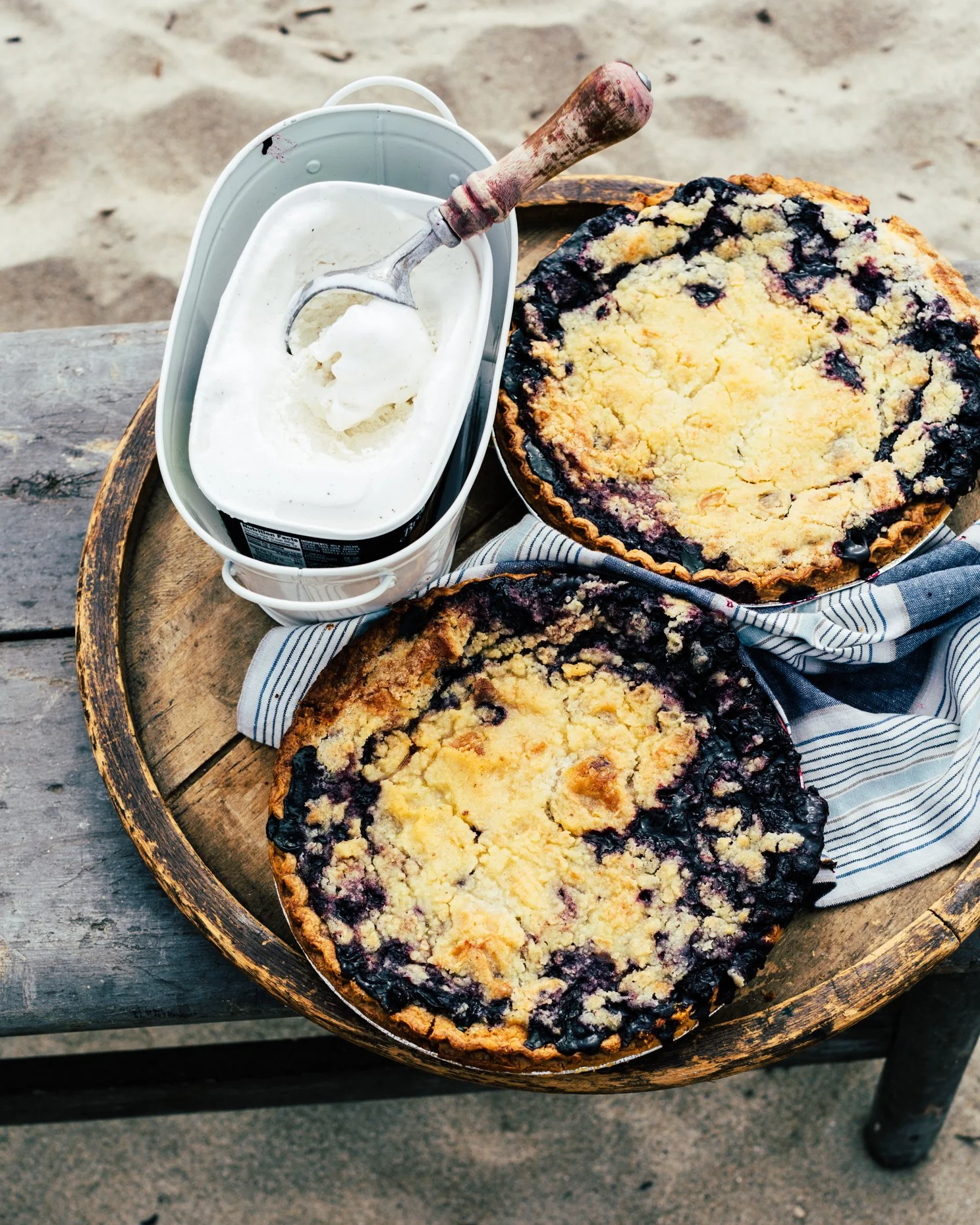 Two blueberry crumb pies with a scoop of vanilla ice cream in a tin container, all placed on a wooden tray lined with a striped cloth on a rustic table.