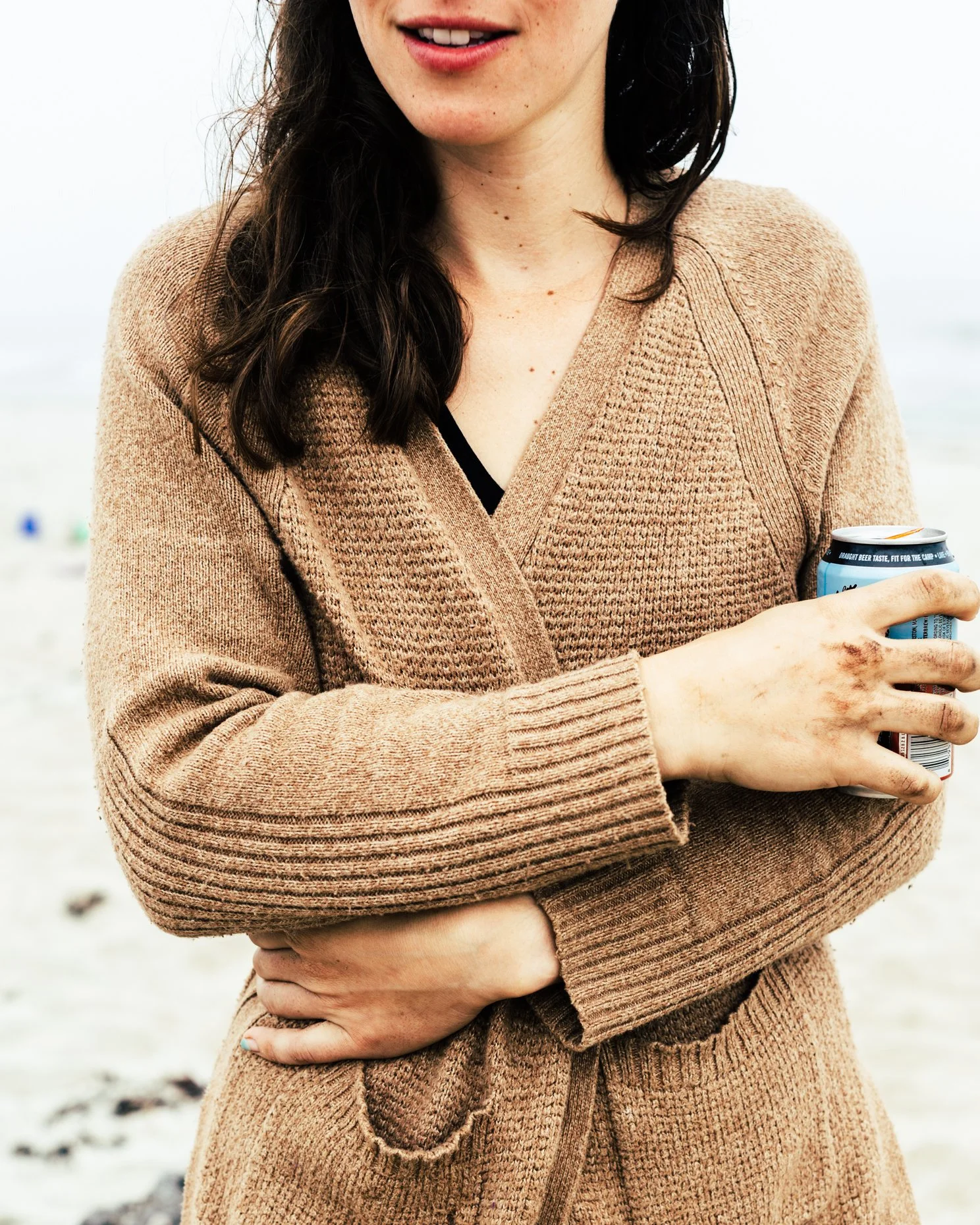 Close-up of a woman wearing a tan knit sweater, holding a can of beverage, standing on a beach.