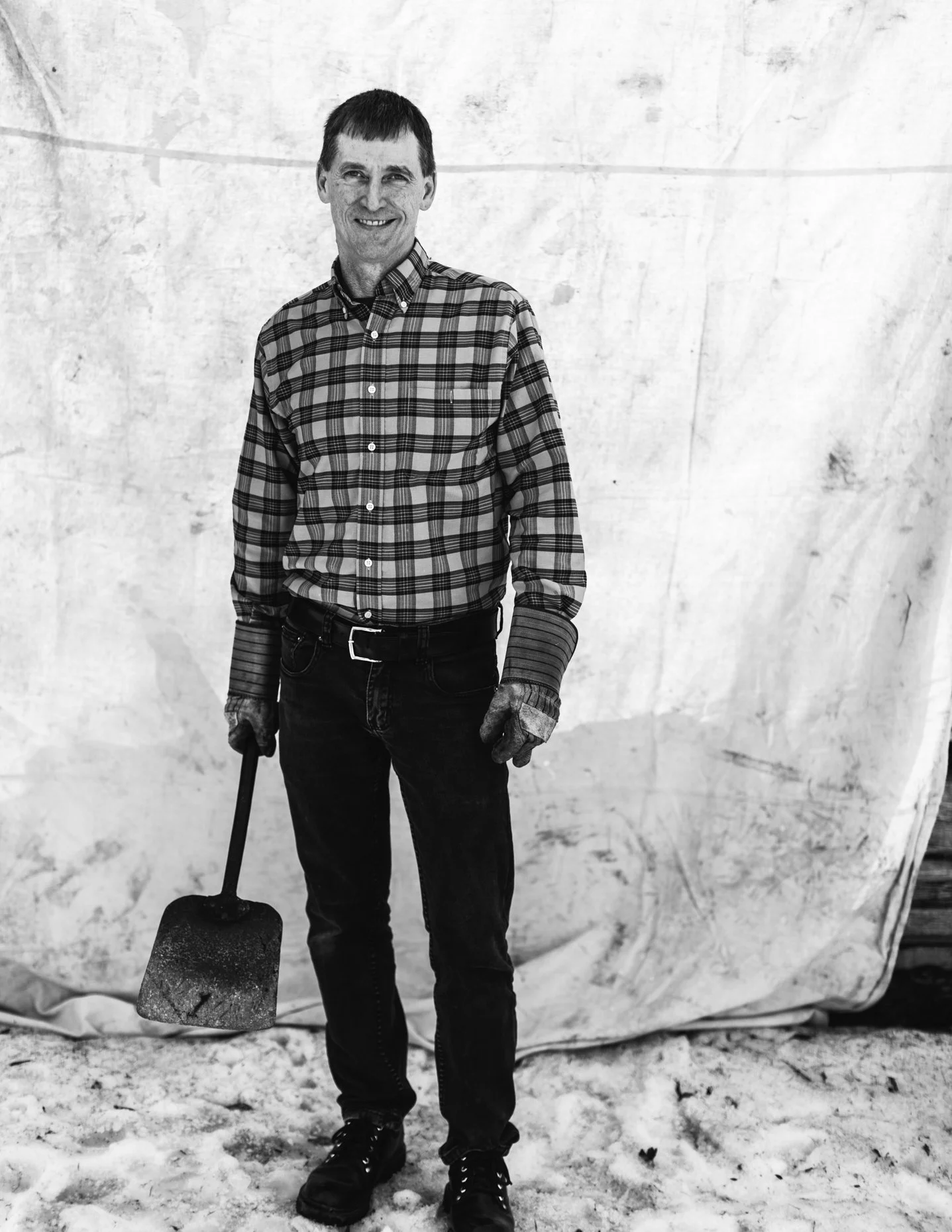 A smiling man in a plaid shirt, jeans, and gloves holding a shovel stands on snow in front of a large, textured white backdrop.