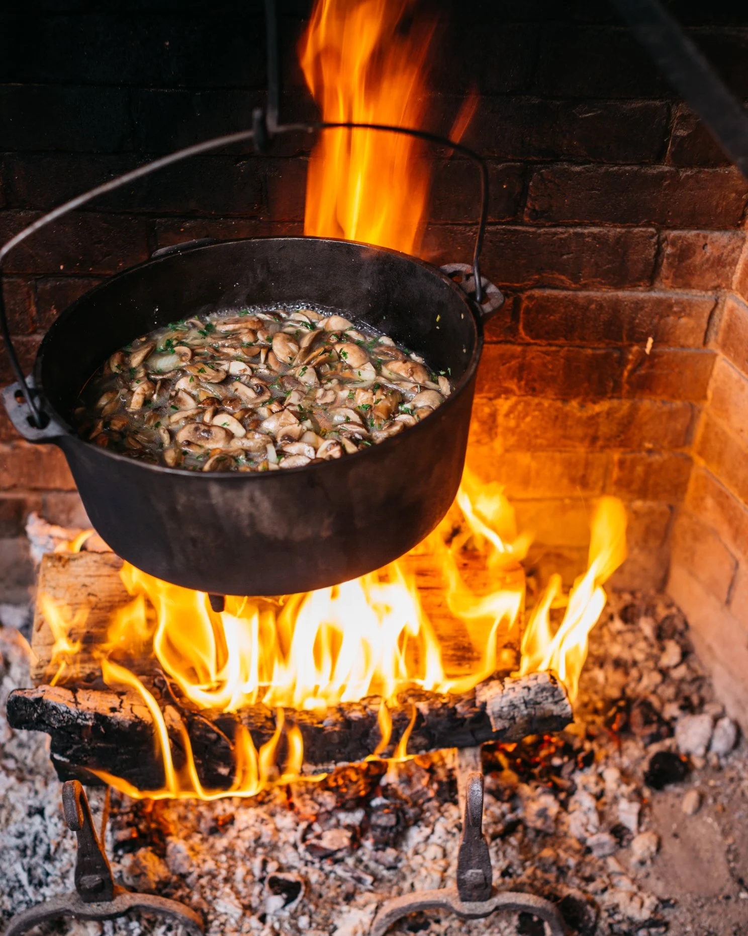 A black pot with chopped mushrooms and herbs cooking over an open wood fire in a brick oven.
