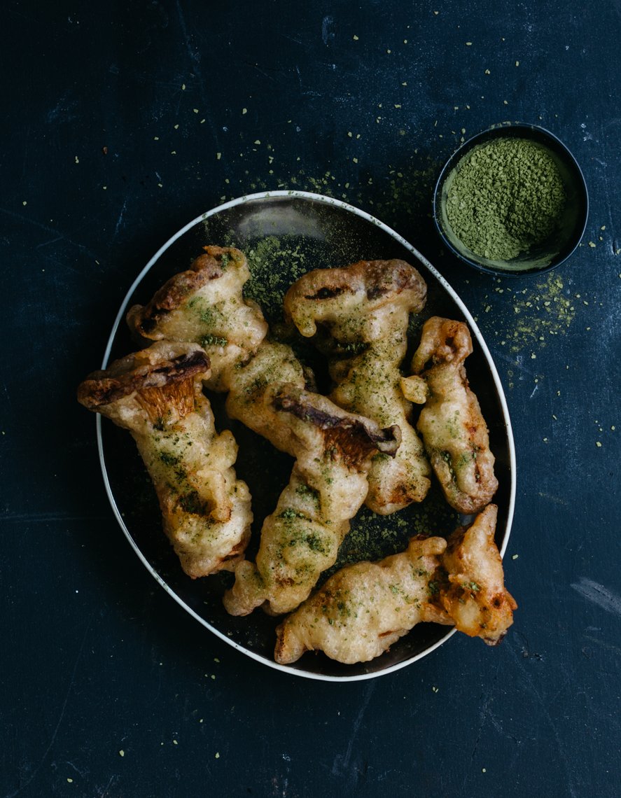 A black plate with deep-fried mushroom tempura garnished with green seasoning, accompanied by a small bowl of matcha green tea powder on a dark surface.