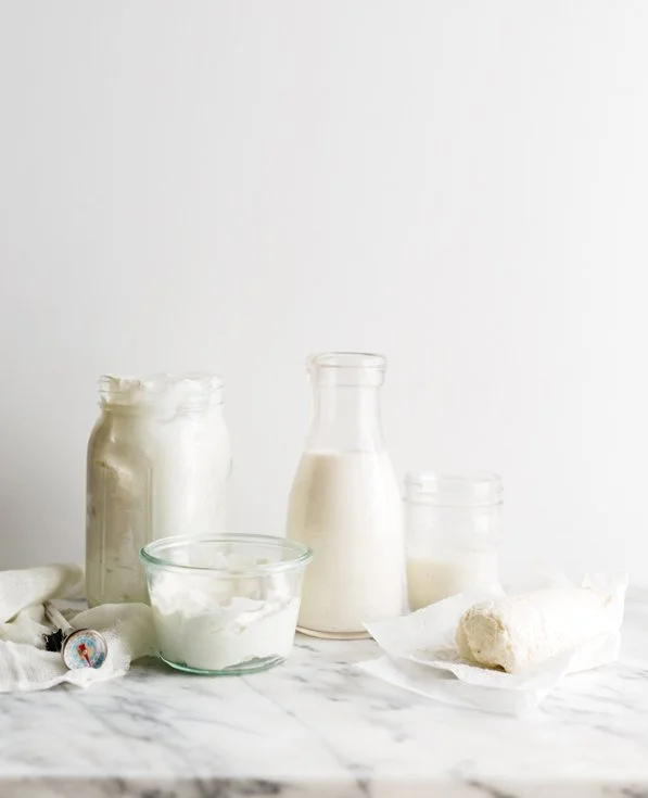 Assorted containers of dairy products, including yogurt, milk, and cheese, on a marble surface with a cheese wheel on a paper napkin.
