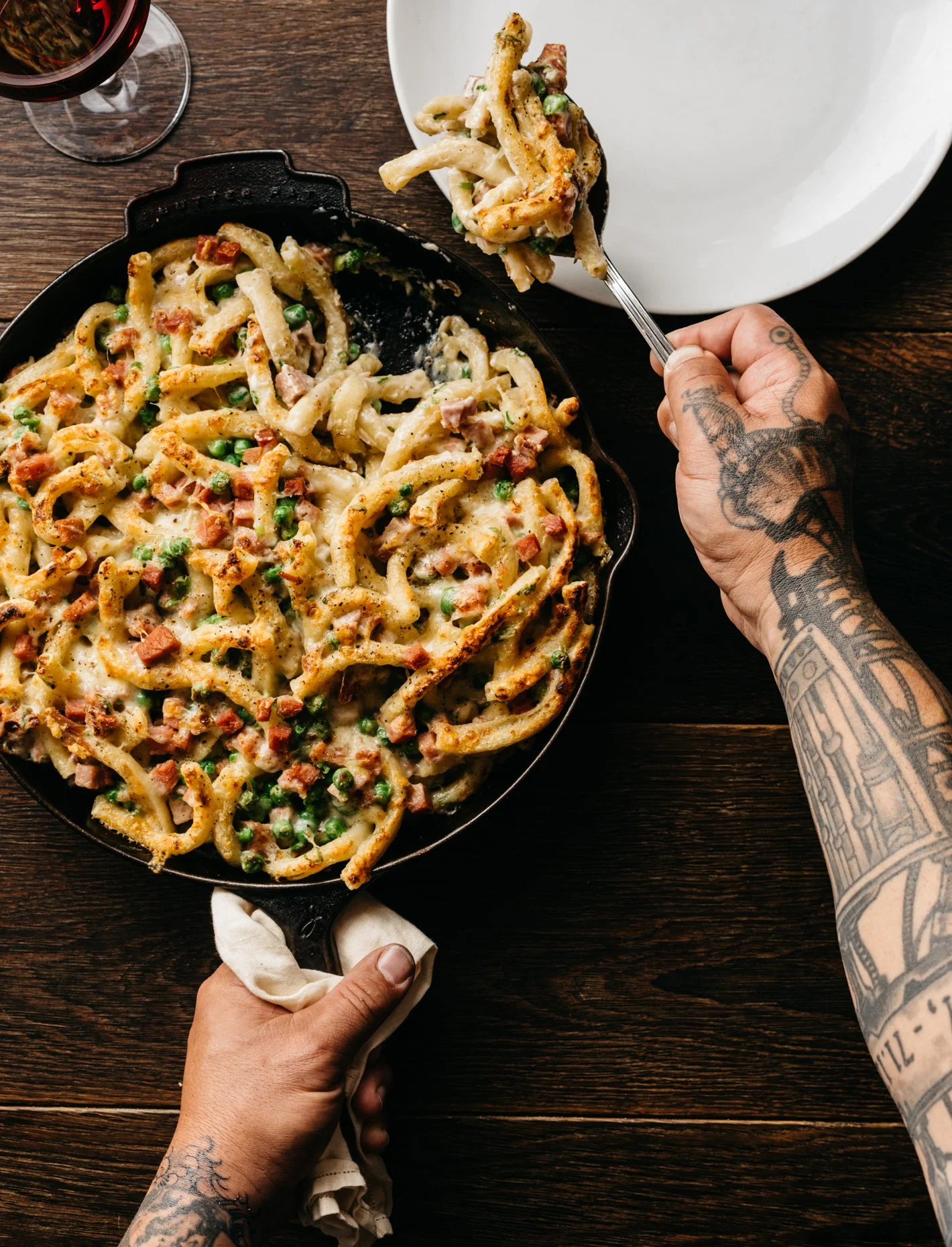 Person serving creamy pasta with peas and ham from a skillet onto a white plate