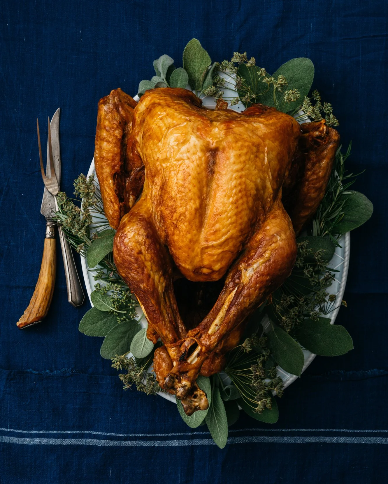 A roasted whole turkey on a white platter garnished with green herbs, placed on a dark blue tablecloth.