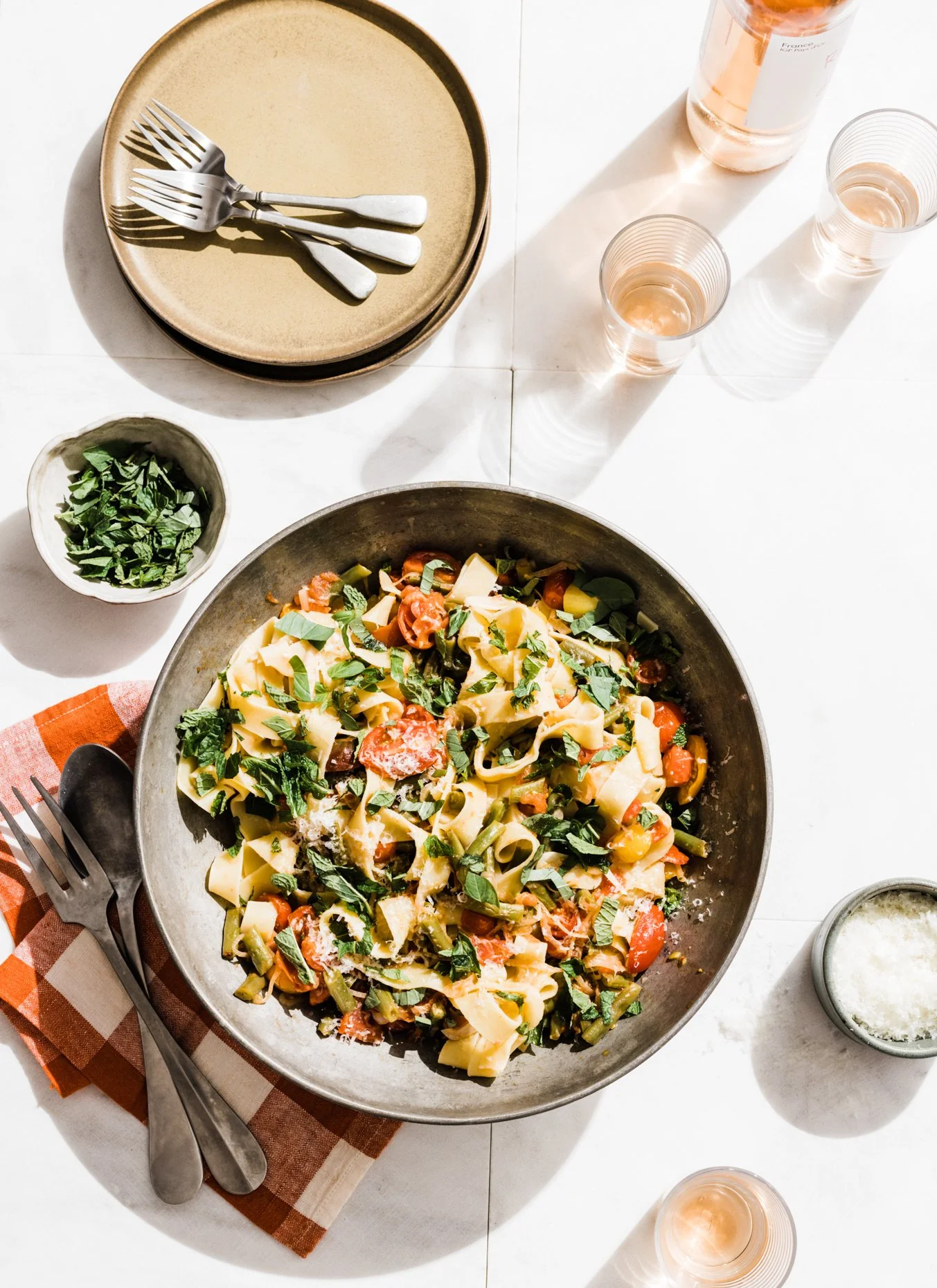 A bowl of pasta with cherry tomatoes, leafy greens, and grated cheese on a white table, with plates, forks, knives, wine glasses, a small bowl of basil, and a bottle of wine.