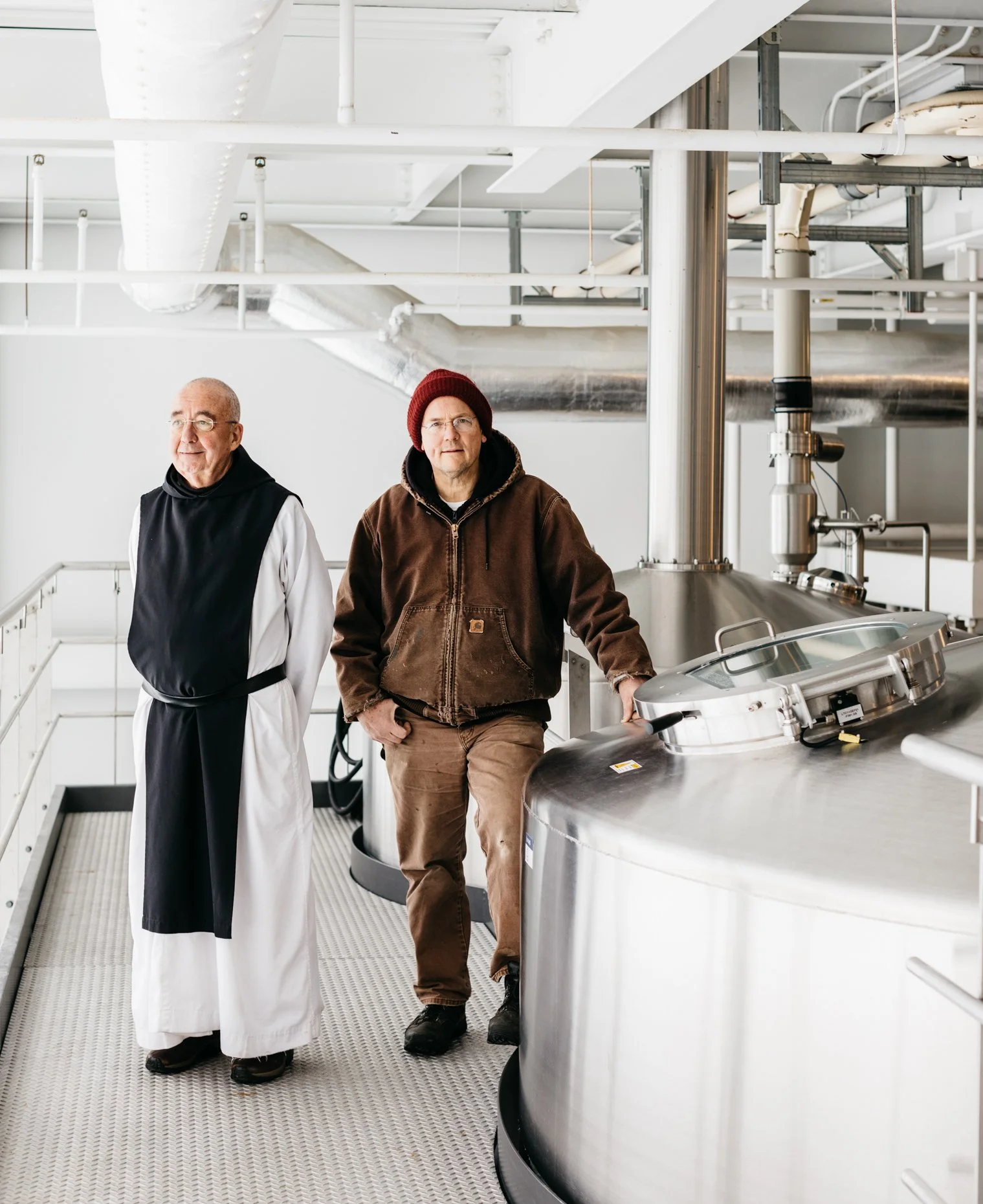 Two men standing in a brewery, one in a black and white clerical robe and the other in a brown jacket and red beanie, next to large stainless steel fermentation tanks.