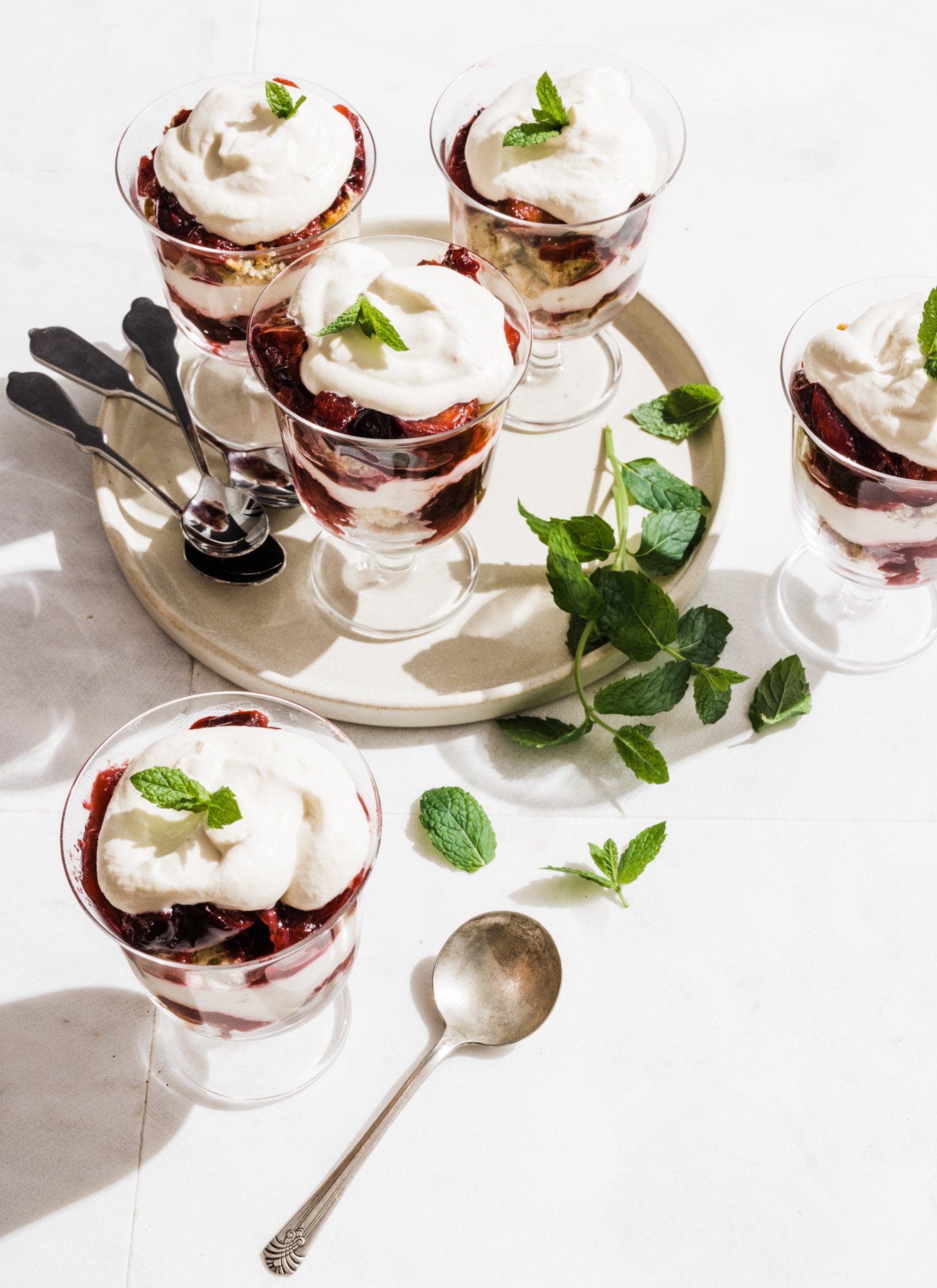 Six dessert glasses filled with berry parfaits topped with whipped cream and mint leaves, arranged on a white surface with scattered mint leaves and a silver spoon.
